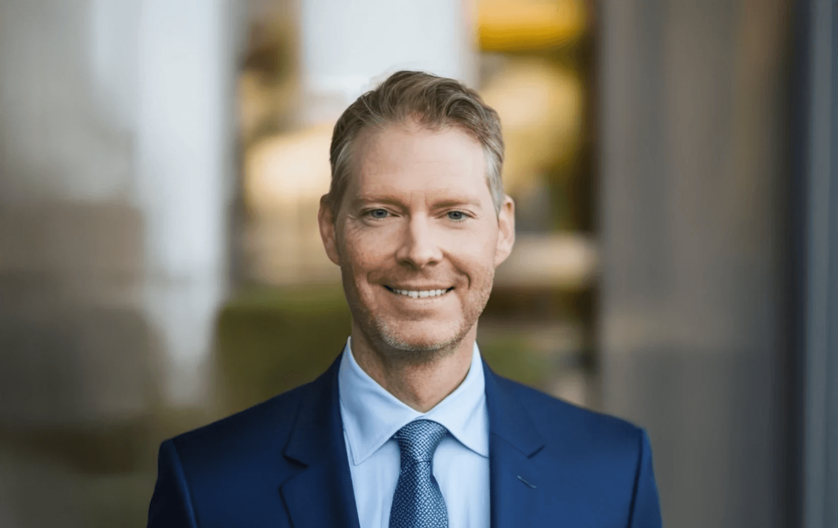 Timo Neustock, Geschäftsführer der ABENA GmbH, Portrait eines Mannes im blauen Anzug und Krawatte vor modernem Bürohintergrund.