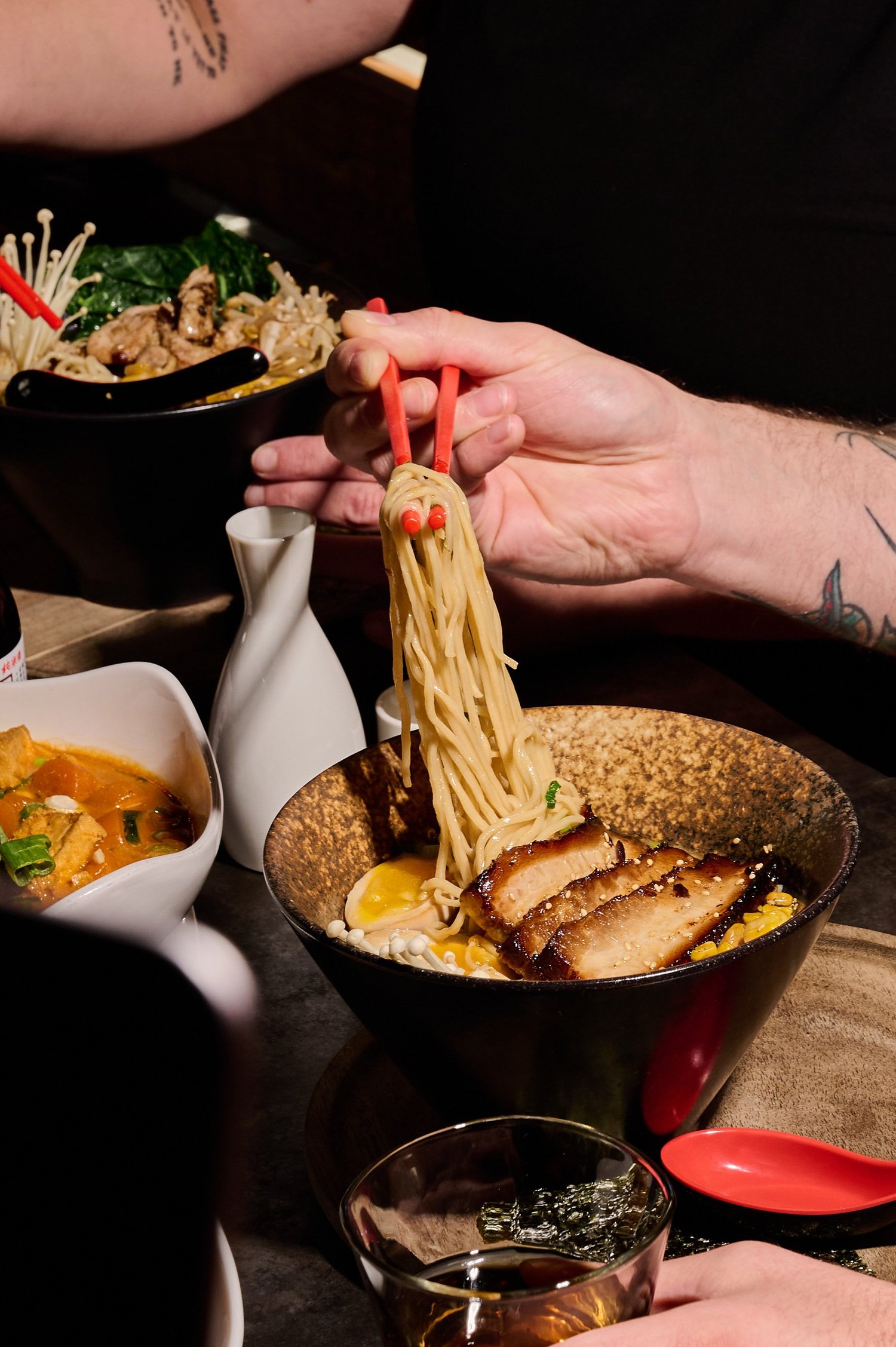 Red chopsticks lifting ramen noodles from a bowl of tonkotsu with chashu pork, more dishes across the table