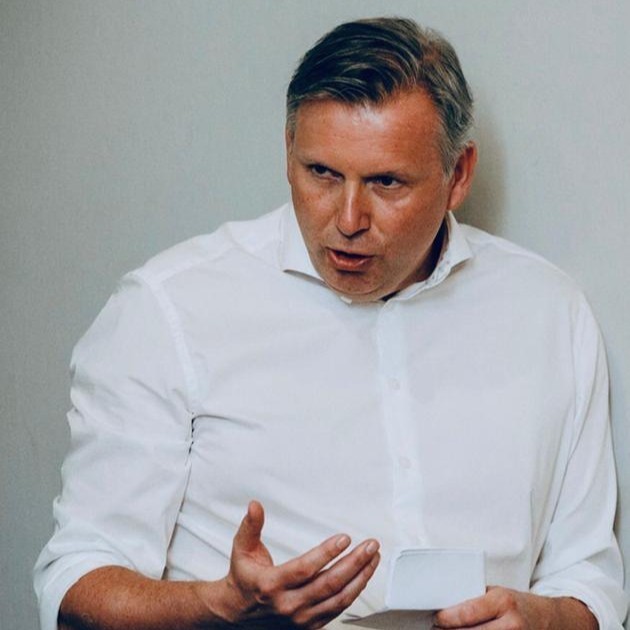 A young woman with long hair smiles while standing against a green backdrop, wearing a casual dark sweater.