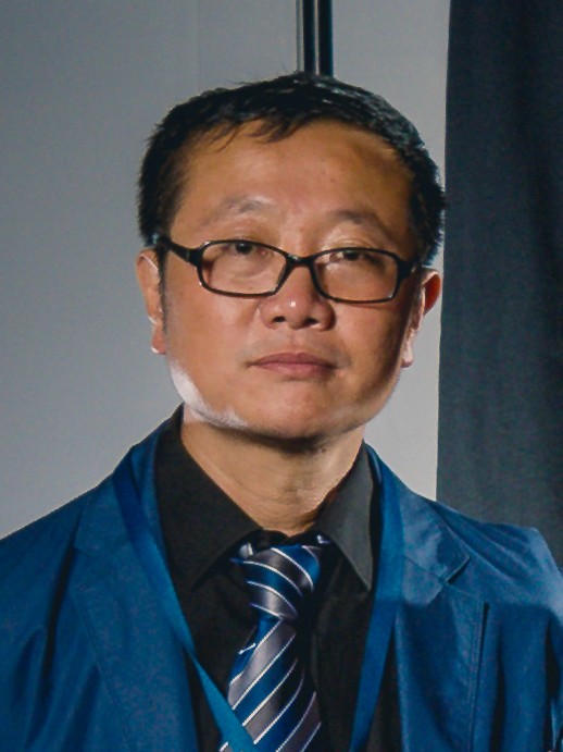 A man of Asian descent wearing glasses, a dark suit, and a striped tie stands in front of a neutral background.