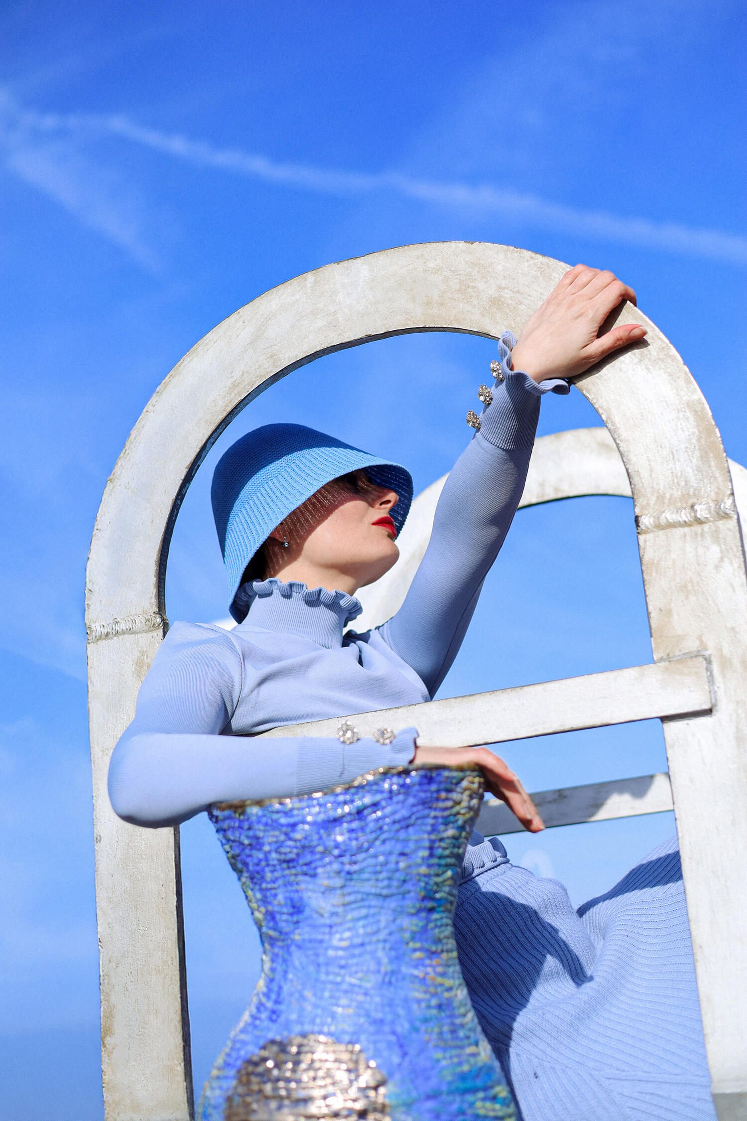 Editorial seaside fashion photography with model posing against bright blue sky and sculptural vase.