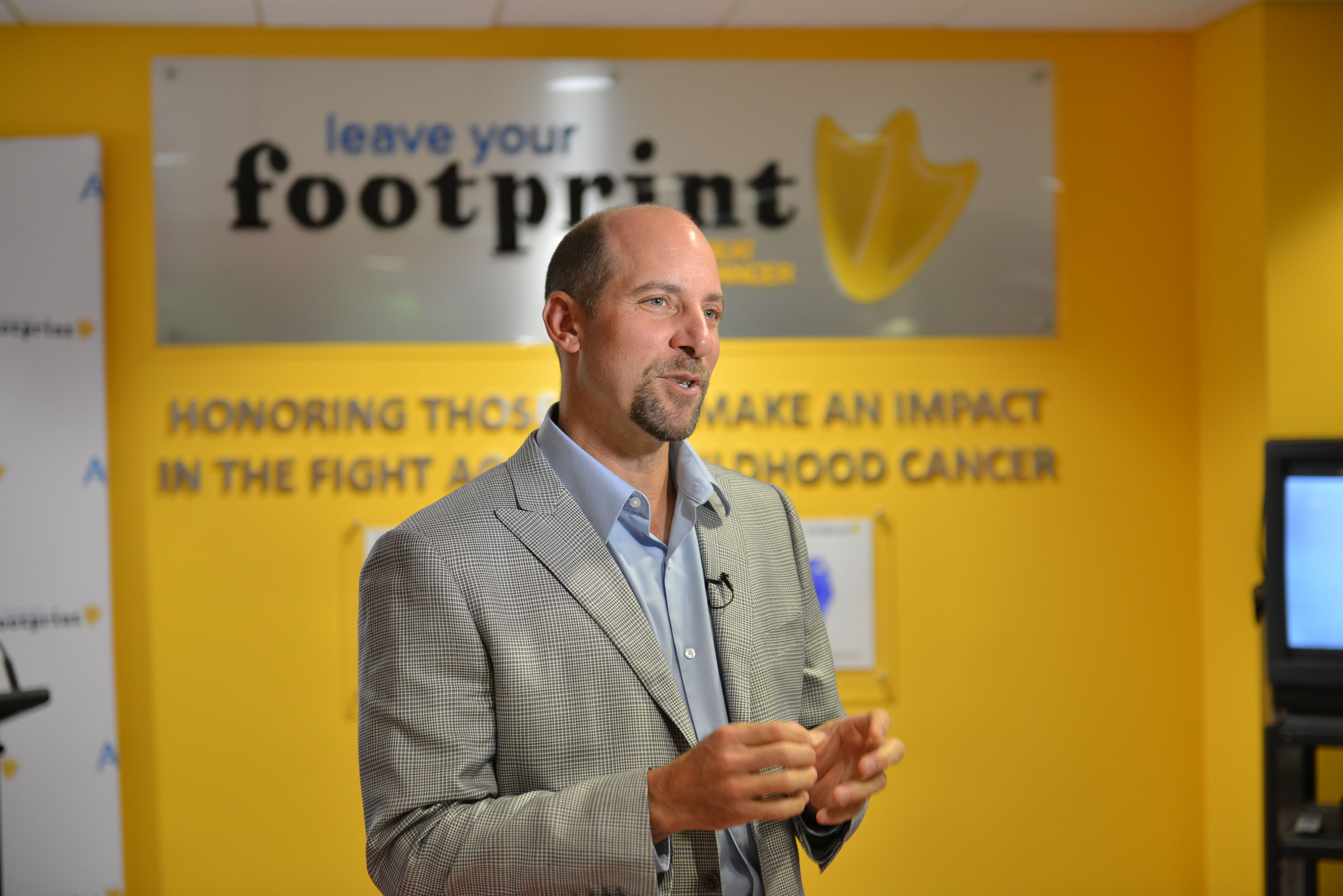 John Smoltz in a gray suit speaks in front of a yellow wall with signs that read leave your footprint and honoring those who make an impact in the fight against childhood cancer.