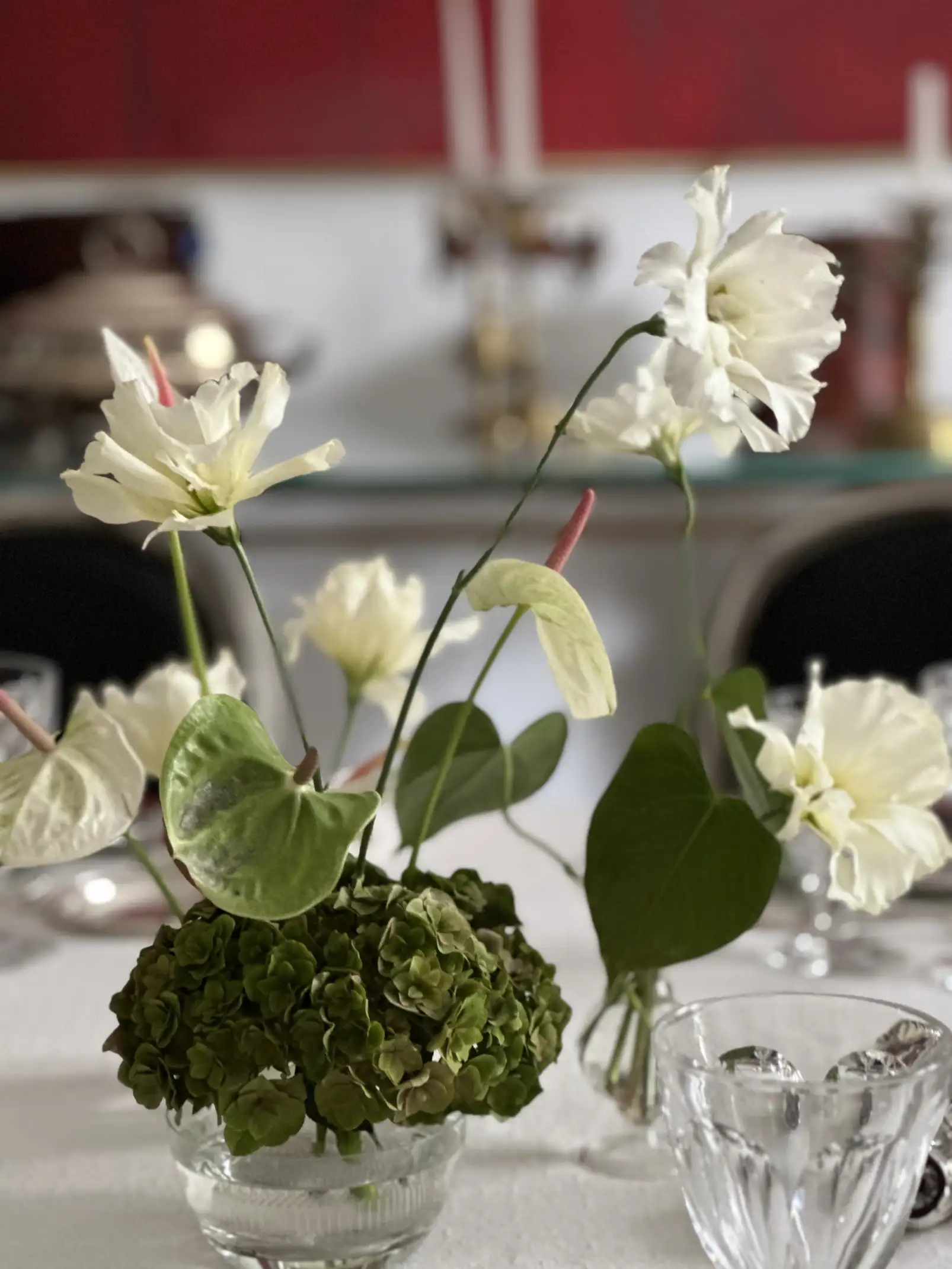 Arreglo floral en tonos blancos y verdes compuesto por anturios pálidos, flores delicadas de pétalos rizados y hojas grandes, dispuesto en un pequeño florero de vidrio sobre una mesa con mantel blanco y copas transparentes.