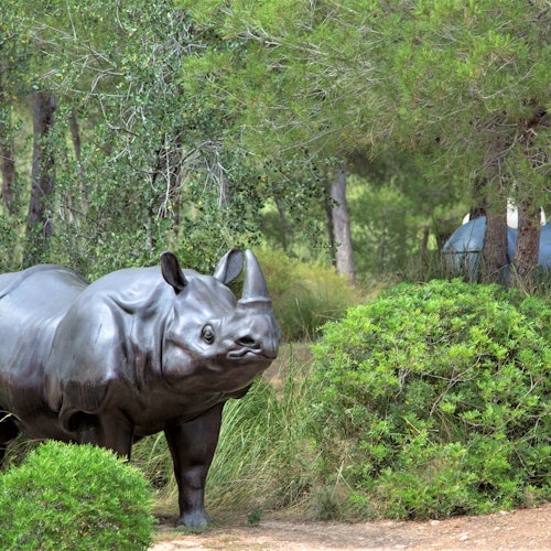 A life-sized rhinoceros statue surrounded by lush greenery and trees in a wooded area.