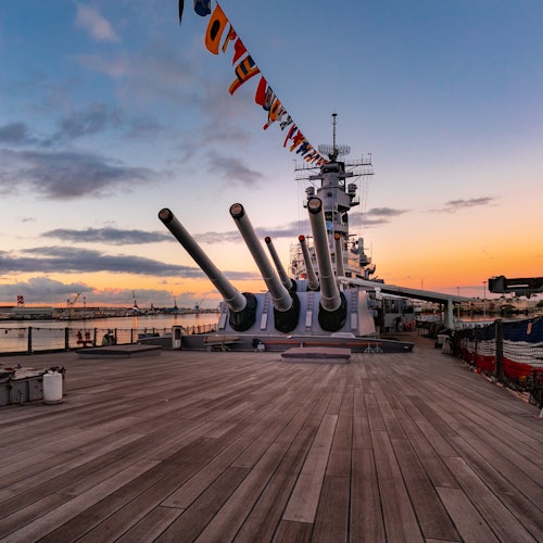 ดาดฟ้าเรือรบยามพระอาทิตย์ตกพร้อมปืนใหญ่สามกระบอกที่เล็งไปข้างหน้า และธงสัญญาณนาวีที่แขวนอยู่ด้านบน