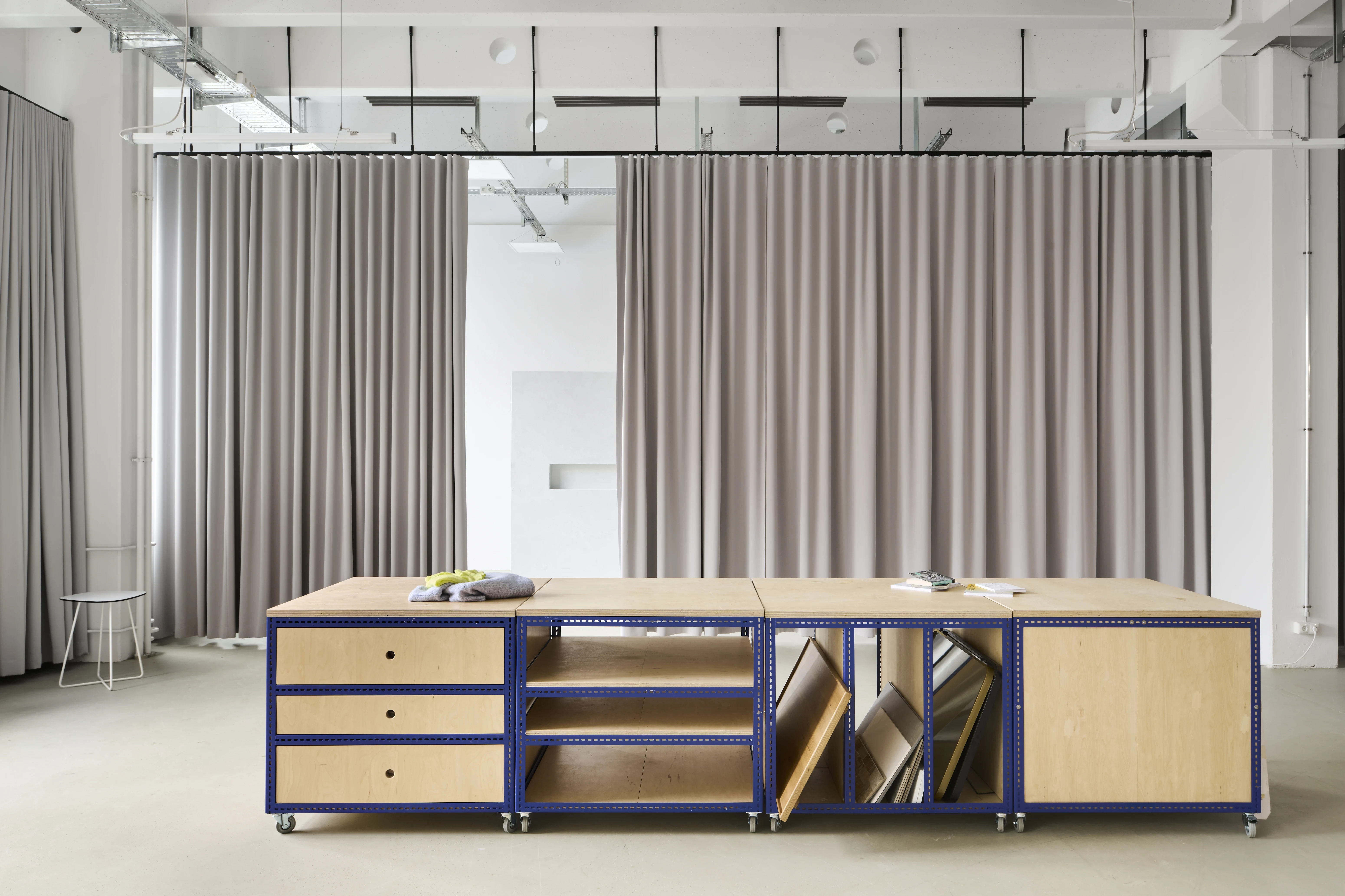 Mobile storage sideboard at the FLACONI CONTENT STUDIO made of birch plywood with blue steel frames, holding drawers, shelves and leaning boards in front of grey curtains.