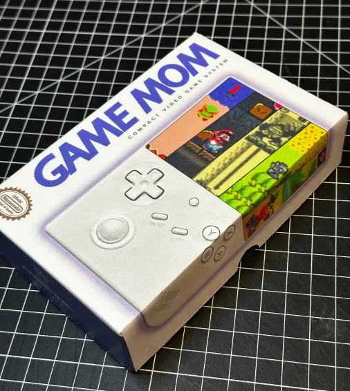 A retro video game console box labeled "GAME MOM" features a design reminiscent of classic consoles, highlighted with a white controller graphic and colorful game scenes, displayed on a black grid background, showcasing unique 'Rob Harrigan Design' elements.