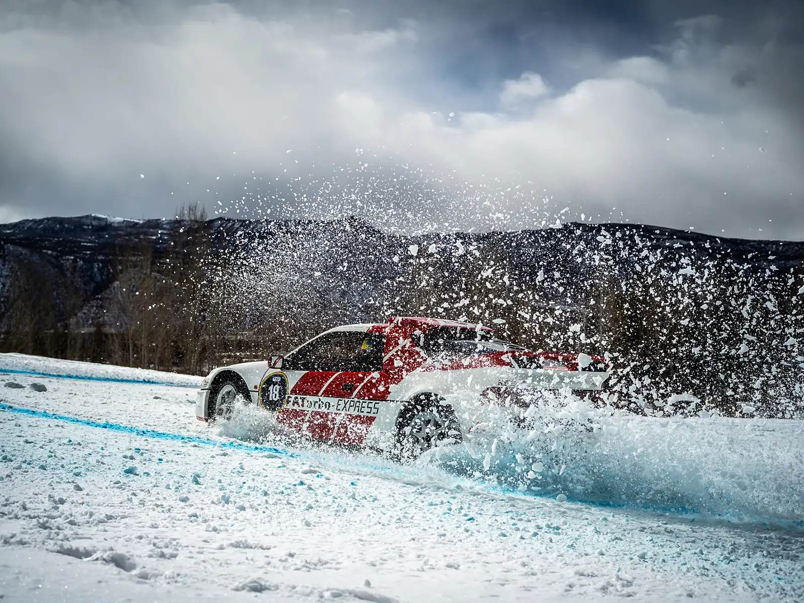 A red and white rally car speeds through a snowy landscape, kicking up snow as it navigates the icy terrain.