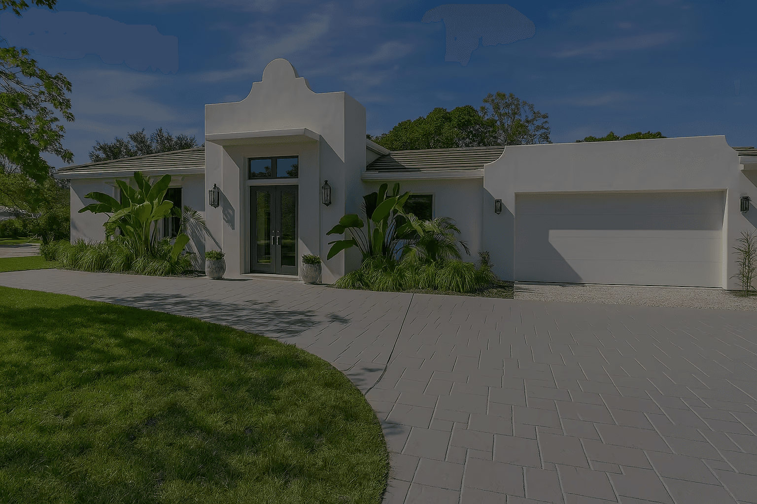 Modern Southwest-style home with clean white stucco exterior and distinctive parapet roofline. Features dramatic front entryway with tall glass doors flanked by lantern sconces, large garage, and minimalist drought-resistant landscaping with tropical accent plants. Gray concrete paver driveway complements the architectural style against deep blue sky