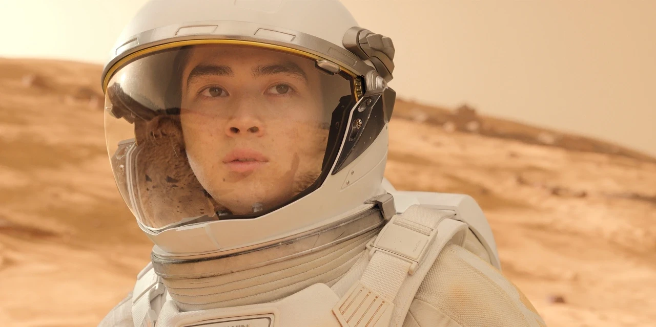 A young man wearing a white space suit and helmet stands on a rocky, Mars-like landscape with a focused expression, highlighting exploration and adventure in a Martian environment.