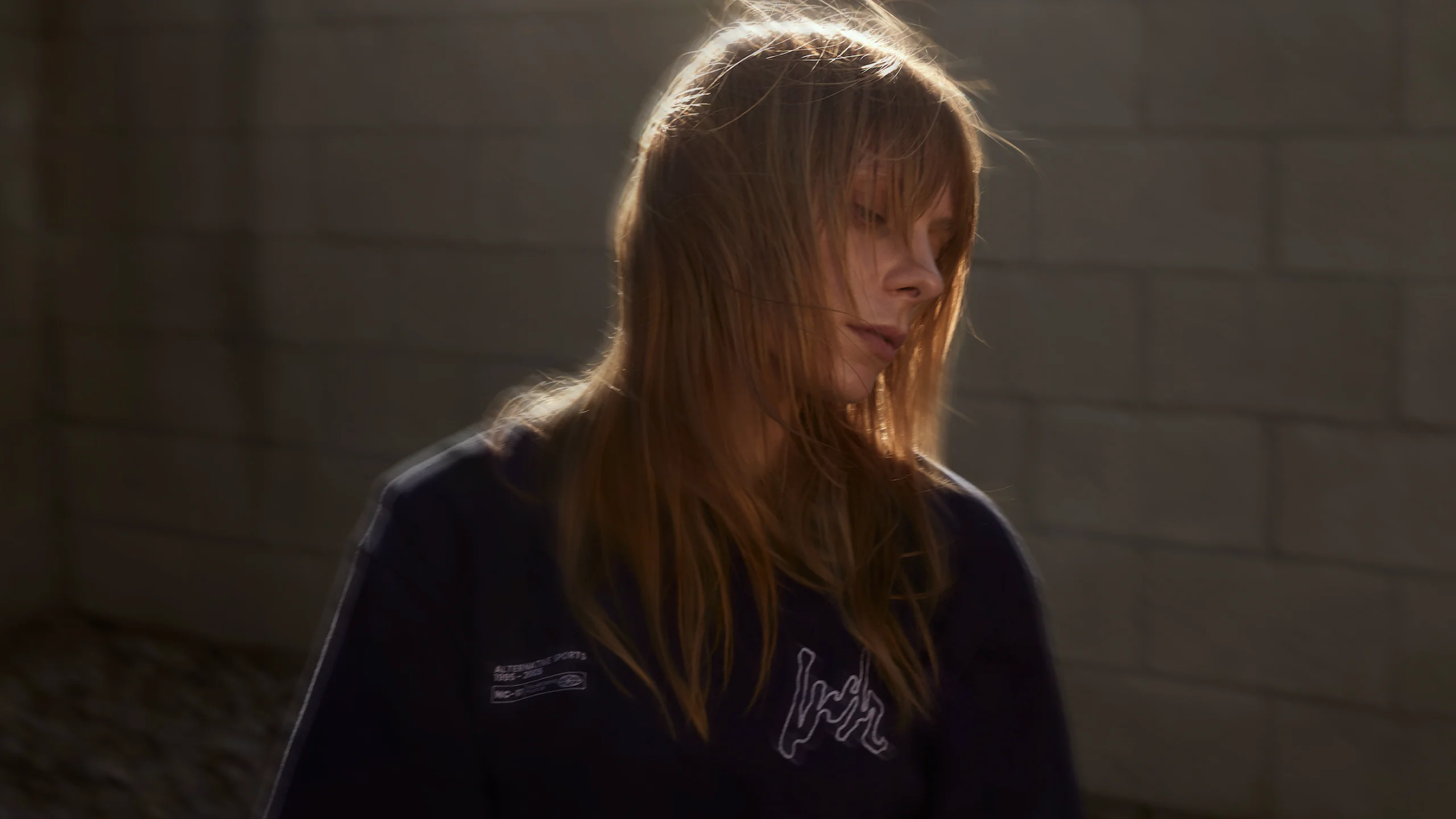 A woman in afternoon light with her hair blowing, wearing the Volume LWH Alternative Sports 02 T-shirt in front of a sunlit cinderblock wall.