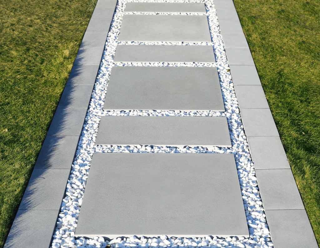 Moderní dům s velkými okny, kamennou cestičkou a upraveným trávníkem, obklopený stromy pod jasnou modrou oblohou.