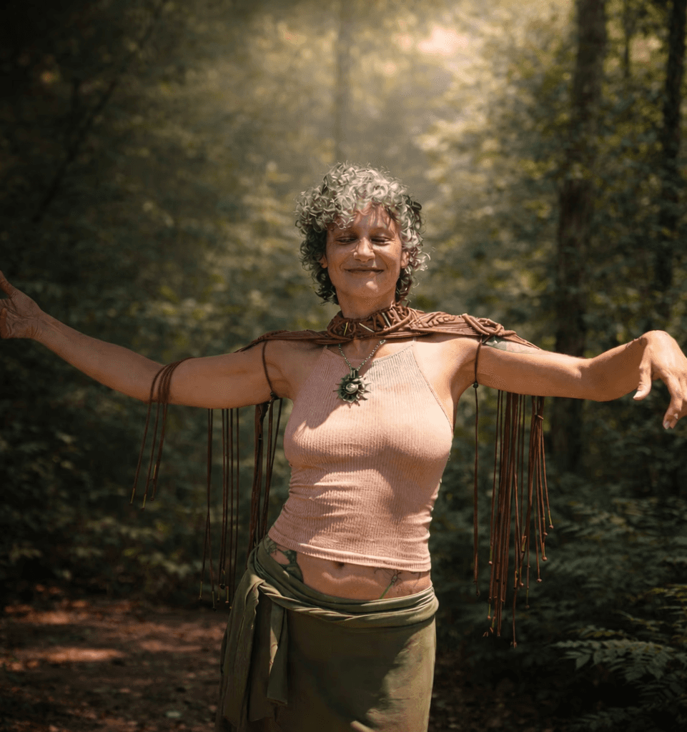 A person with short hair stands in a forest, arms outstretched, surrounded by trees and soft light.
