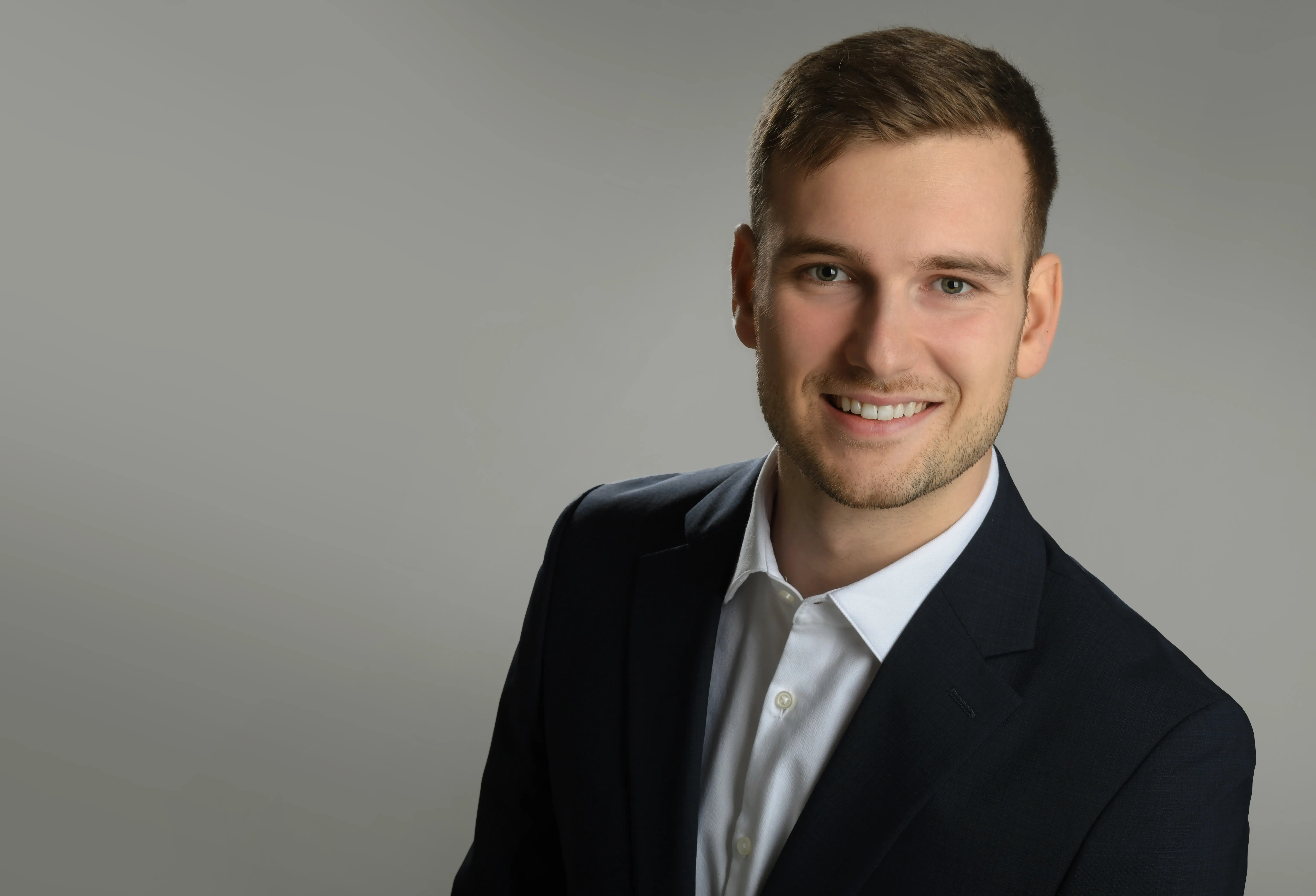A smiling man with curly hair is sitting at a desk, looking at the camera, with a laptop and a lamp in the background.