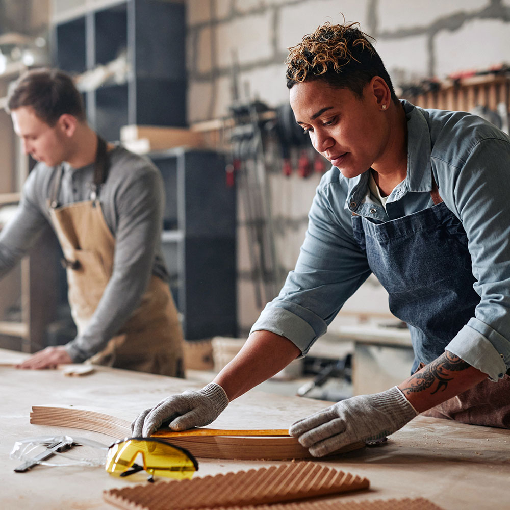 A woman wearing a denim apron and work gloves carefully measures a wooden plank with a tape measure in a carpentry workshop. She is focused on her task at a workbench scattered with wood pieces and tools, including a yellow tape measure. A colleague works in the blurred background, with tools hanging on a pegboard wall behind them, depicting a collaborative skilled trades environment.