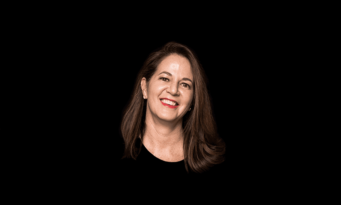 A woman with shoulder-length brown hair is centered against a solid black background. She is smiling and looking directly at the camera. Her skin tone is light, and she wears a dark-colored top. The lighting highlights her facial features and the texture of her hair. No other objects or text are visible in the image.