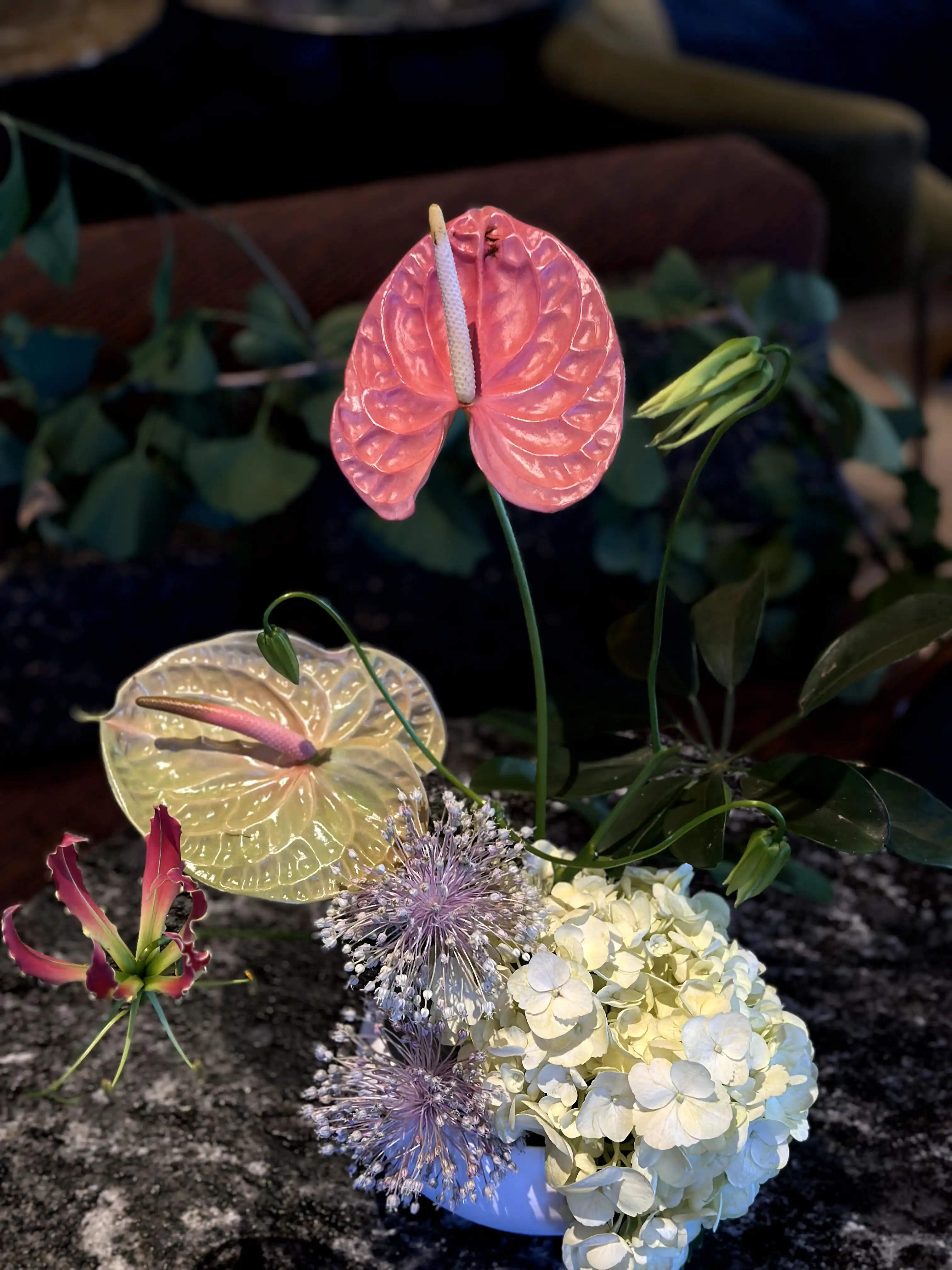 Arreglo floral elegante con anturios rosados y amarillos, esferas de allium lila, hortensias blancas y hojas verdes, dispuesto sobre una mesa de mármol oscuro en un ambiente sofisticado.