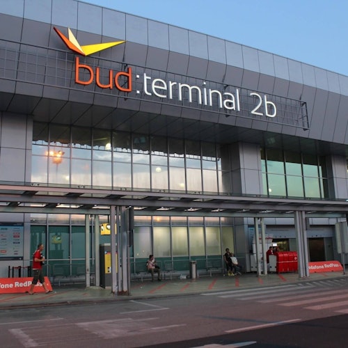 Esterno del Terminal 2b dell'Aeroporto di Budapest con segnaletica, finestre di vetro e diverse persone sedute o in piedi vicino all'ingresso.