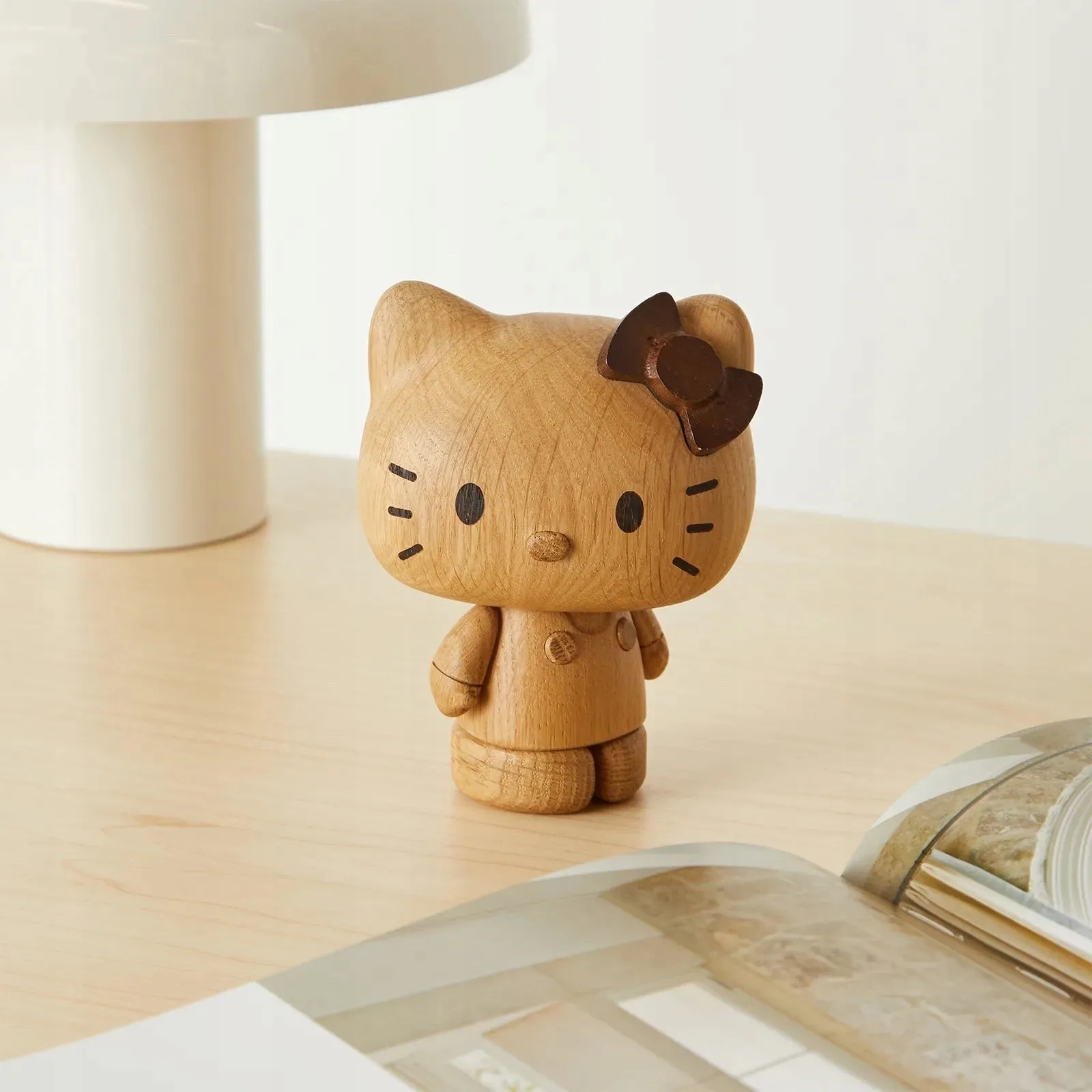 A small, brown wooden hello kitty figurine with a bow, standing on a light wooden desk next to a lamp and notebook.