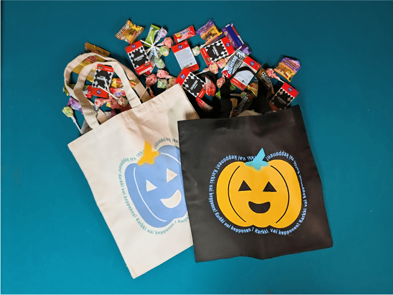 Two tote bags with a mix of American and Finnish candy spilling out. One bag is black with an orange pumpkin jack-o'-lantern, the other is beige with a blue pumpkin jack-o'-lantern. Both pumpkins have the words "Karkki vai Kepponen! repeated outlining the pumpkins. 