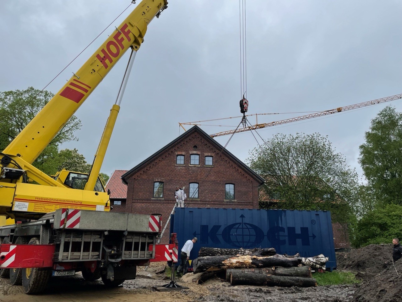 Blauer Seecontainer wird von Kran in der Nähe eines Backsteinhauses angehoben. Koch Logistics