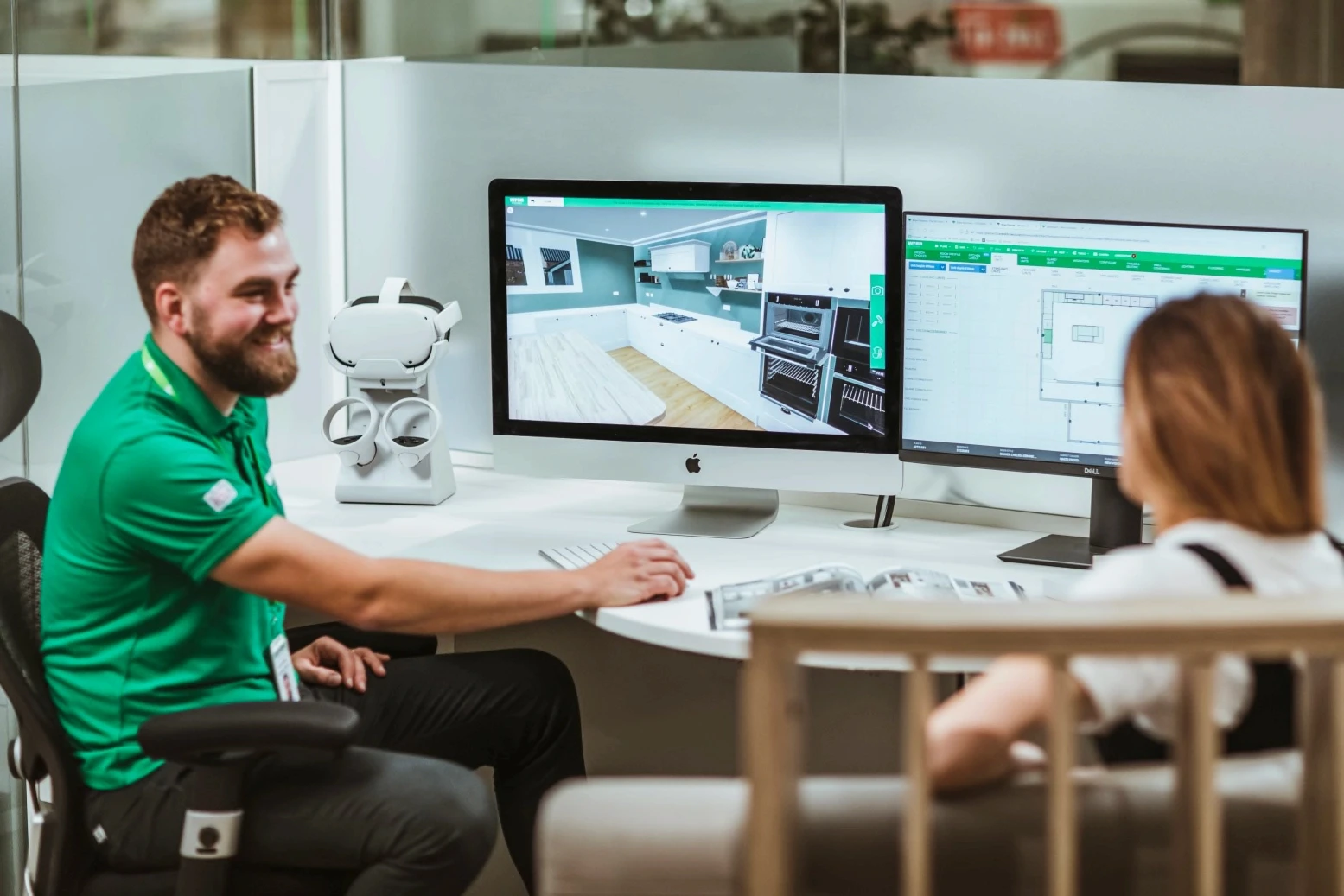 Sales consultant and customer discussing a 3D kitchen design on a computer screen, with a second monitor displaying floor plans.