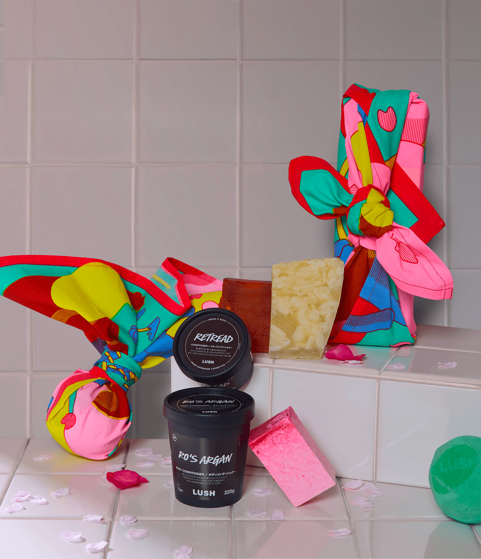Grouped arrangement of Lush bath products, knot-wrapped items, and illustrated fabric on white tiles, with scattered petals and packaging elements.