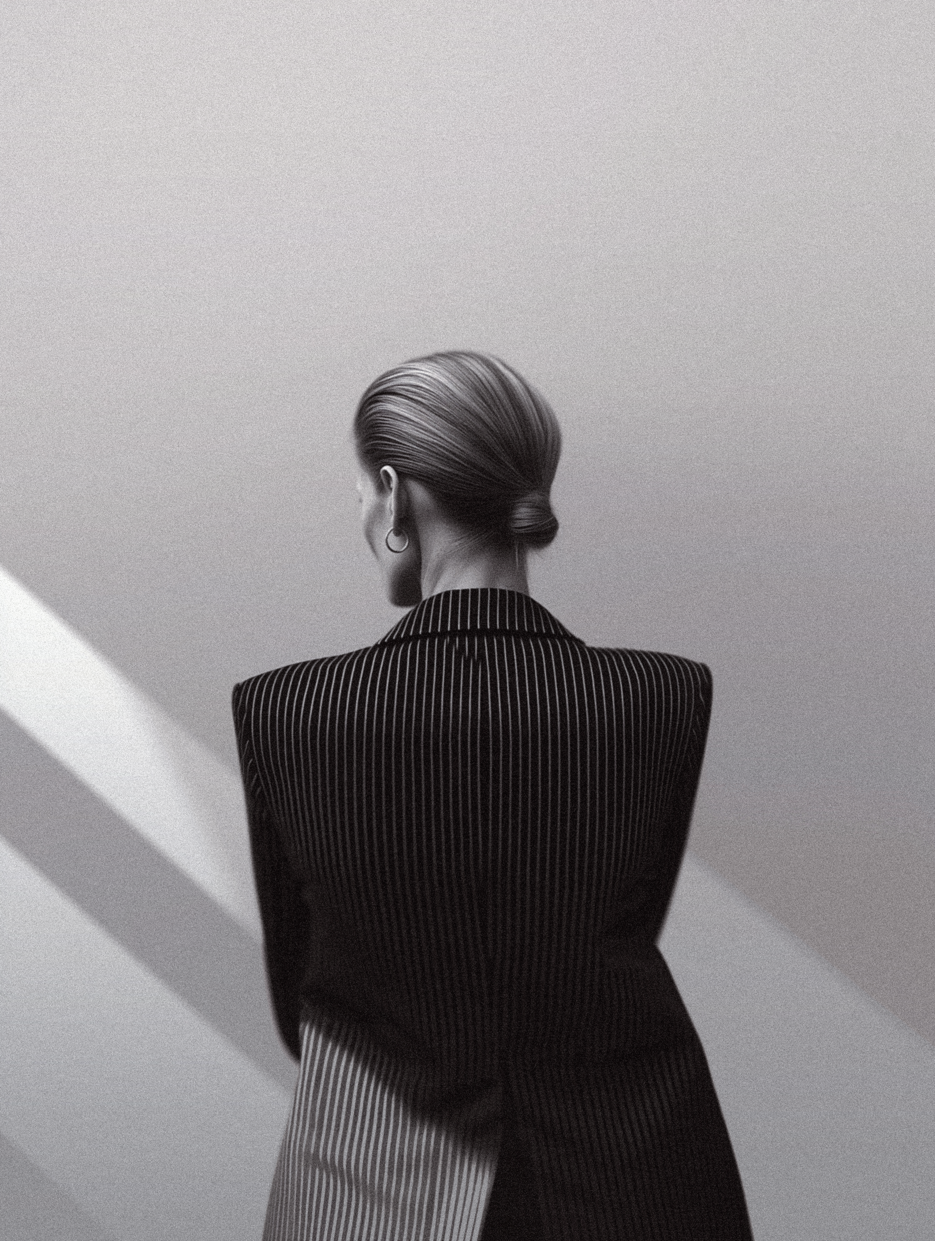 Black-and-white editorial portrait of a woman seen from the back, wearing a structured pinstripe blazer and small hoop earrings.