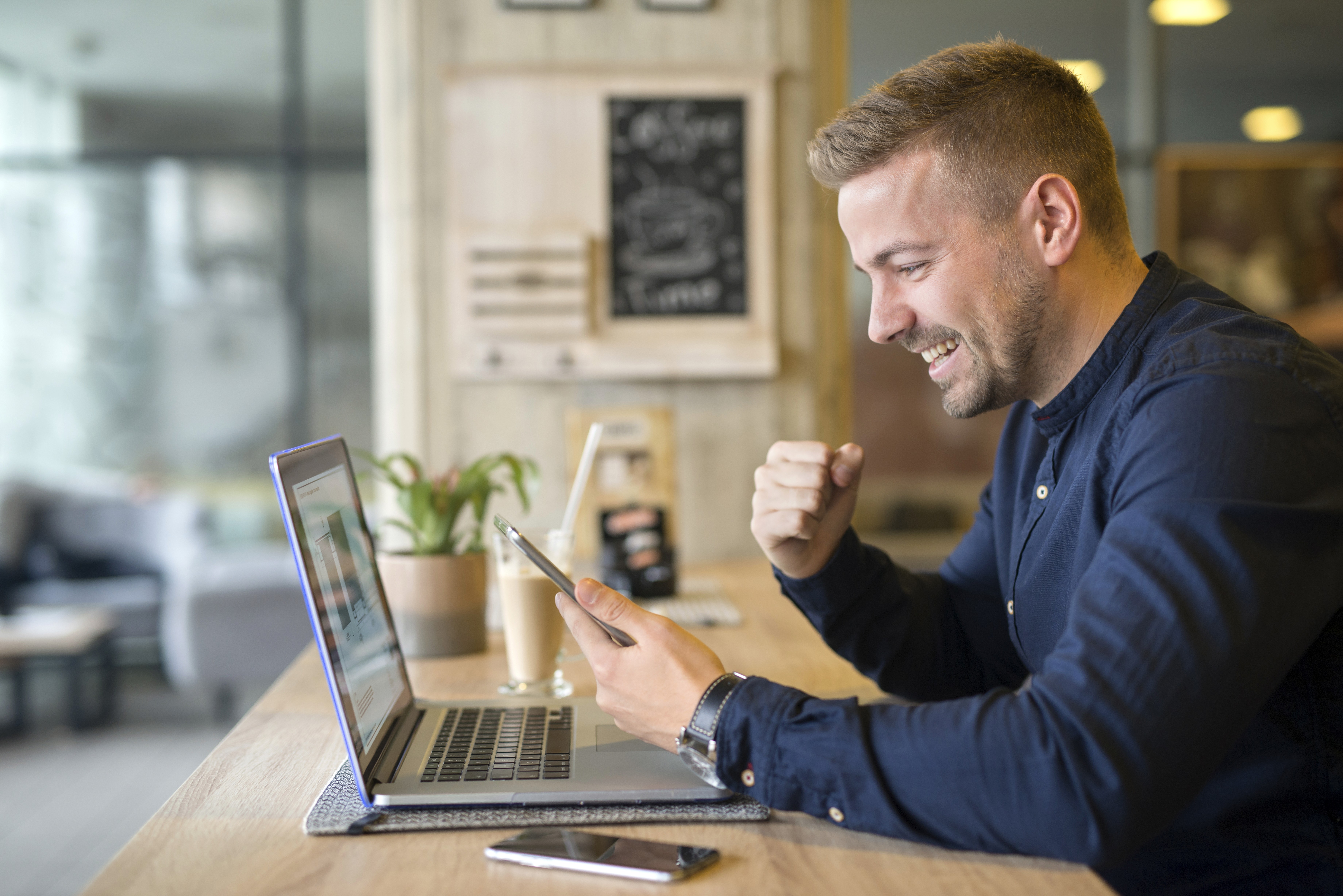 Homem sentado em frente ao notebook, com um celular na mão e comemorando algo que deu certo.
