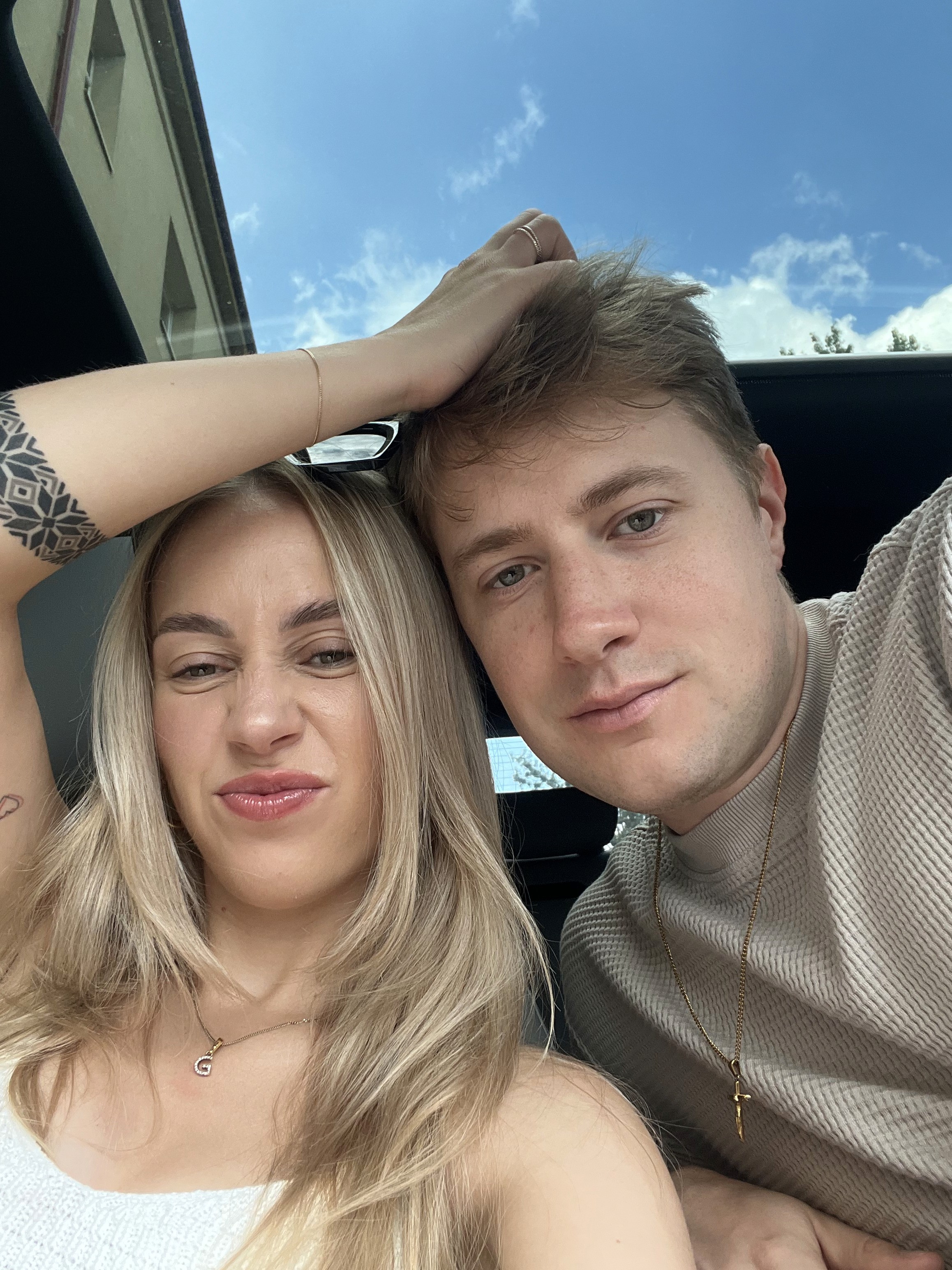 A couple stands on a sandy hill at sunset, facing away, with arms raised in a relaxed "hang loose" gesture. The scene conveys tranquility and joy.