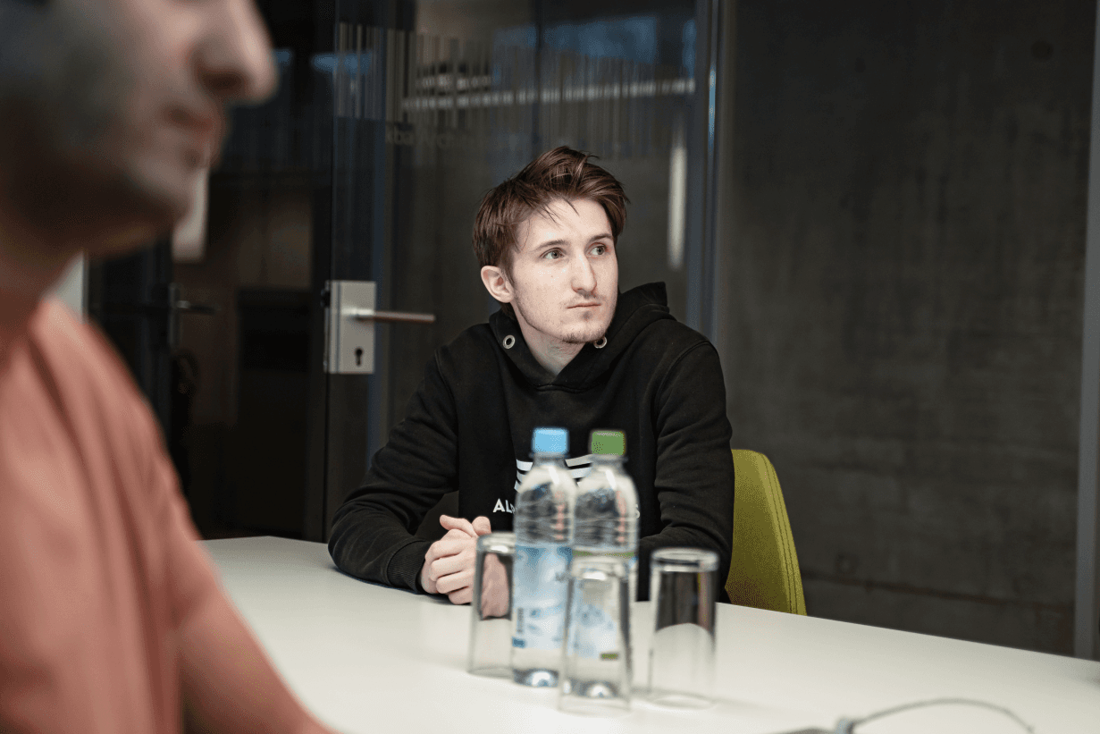 Junger Mann in schwarzem Hoodie sitzt aufmerksam in einem modernen Besprechungsraum der kba Architekten und Ingenieure GmbH; im Vordergrund stehen Wasserflaschen und Gläser auf dem Tisch.