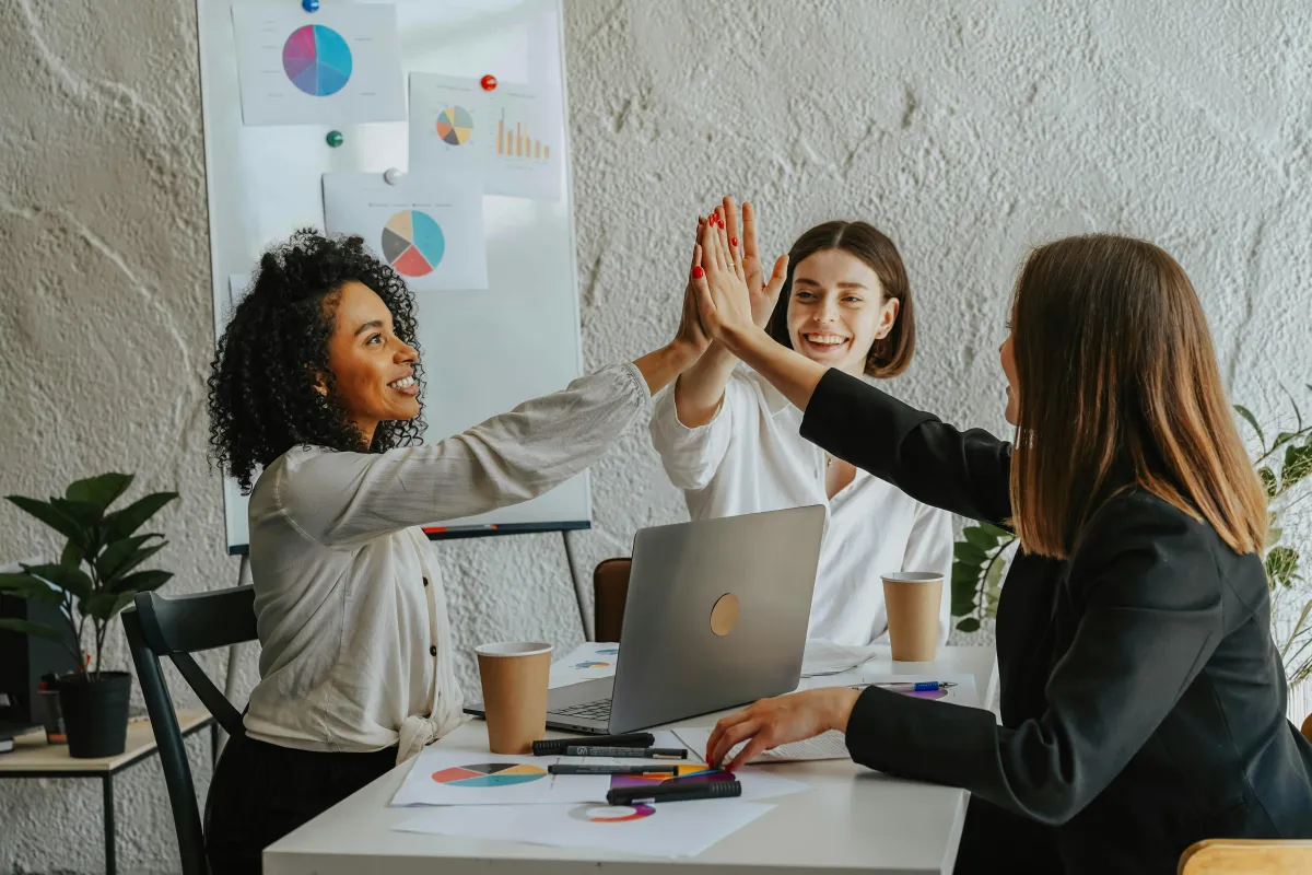 Three diverse women in business attire high-fiving at a desk with charts and a laptop, celebrating financial success and debt freedom with Key 2 Debt Free.