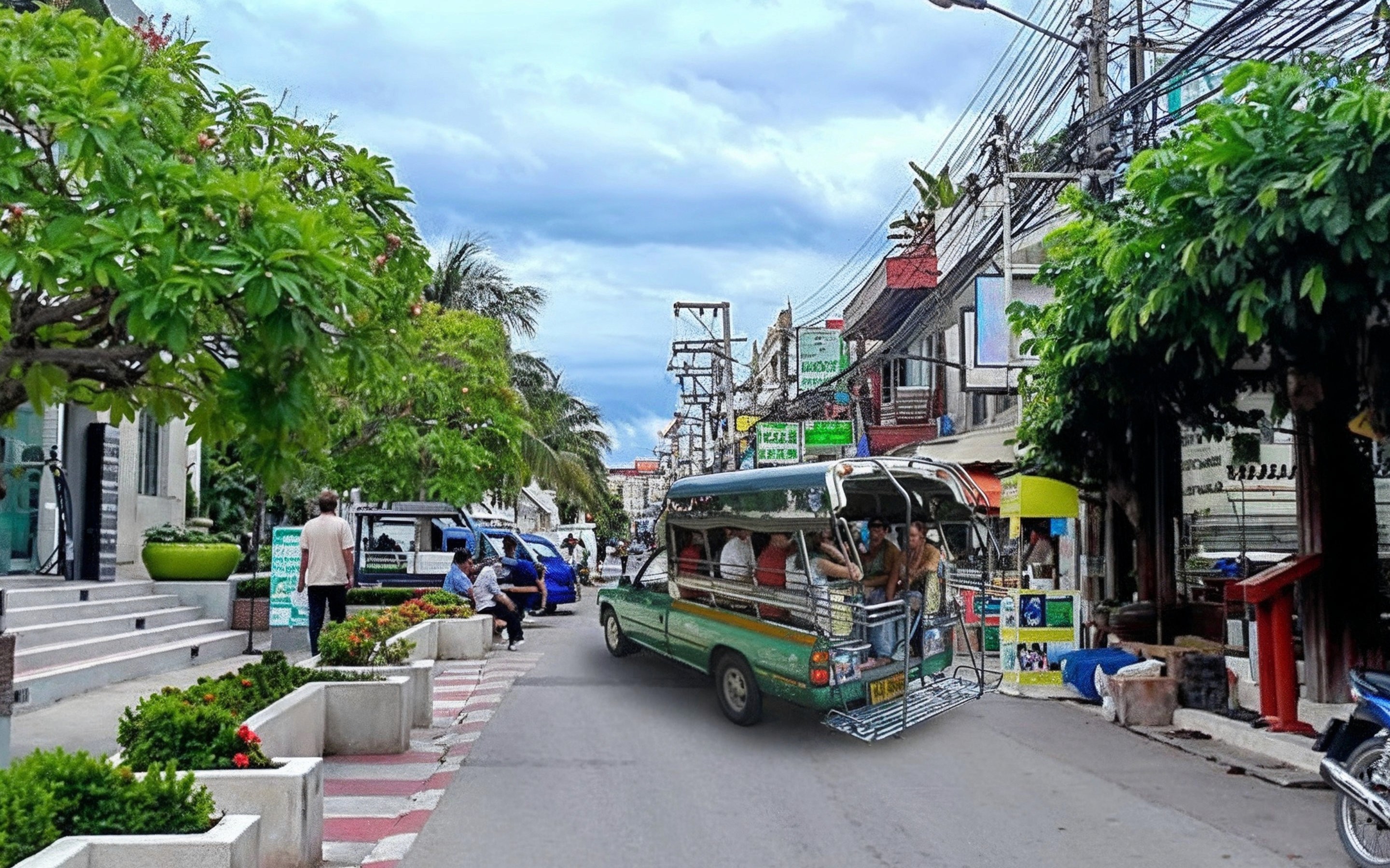 Green Songthaew (Baht Bus) driving down a narrow Hua Hin street lined with shops, trees, and power lines under a cloudy sky.