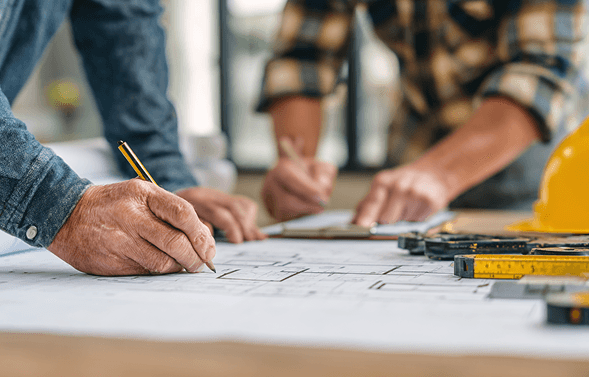 A person wearing a beanie and glasses is working at a computer desk in a sunlit apartment, surrounded by plants, with a cityscape visible through the large windows.