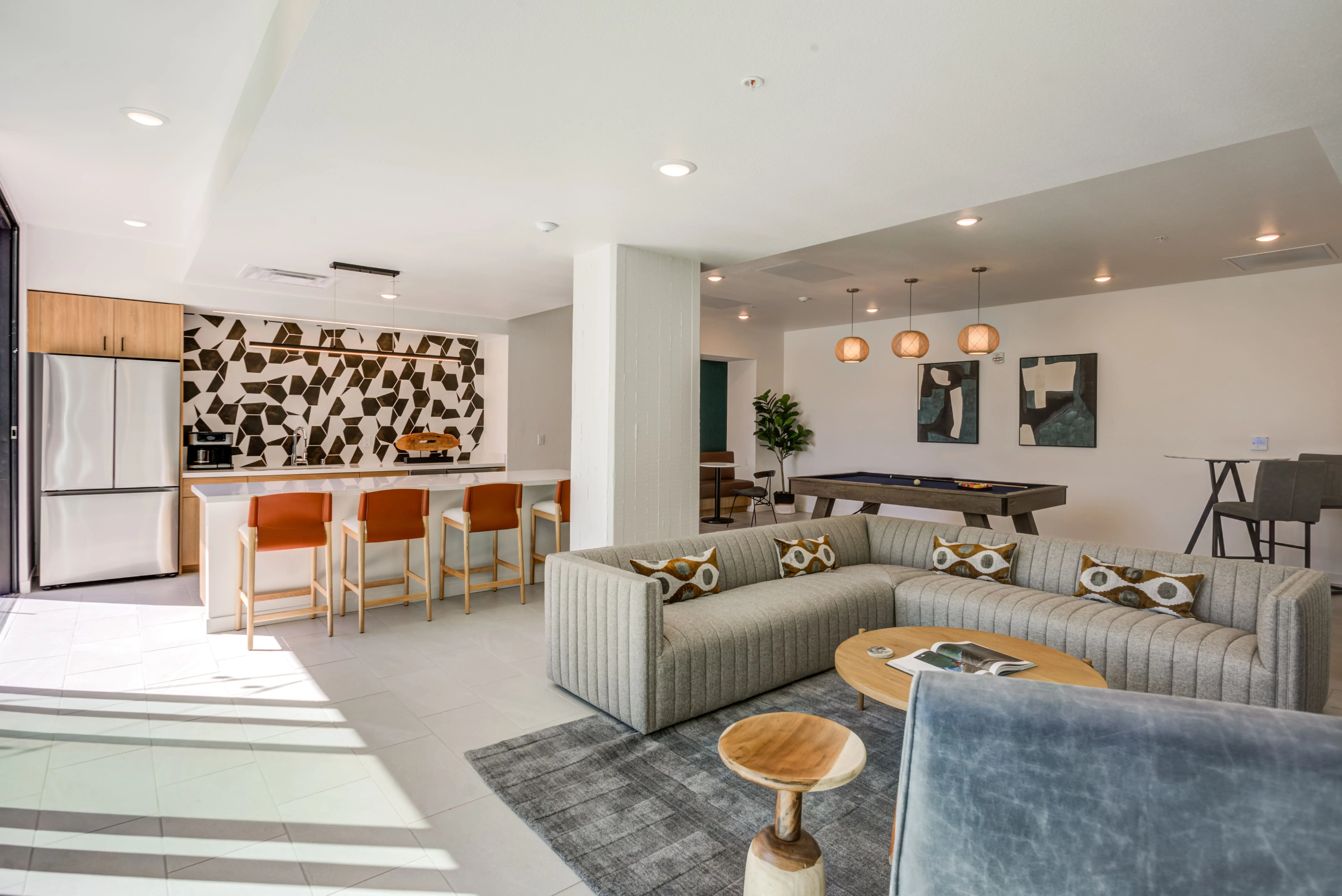 A sun-drenched, contemporary living space featuring a U-shaped gray sectional sofa with patterned cushions, a wooden coffee table, and modern decor. The open kitchen in the background has a mosaic backsplash, stainless steel appliances, and an island with orange barstools—epitomizing modern living spaces in Midtown Sacramento.