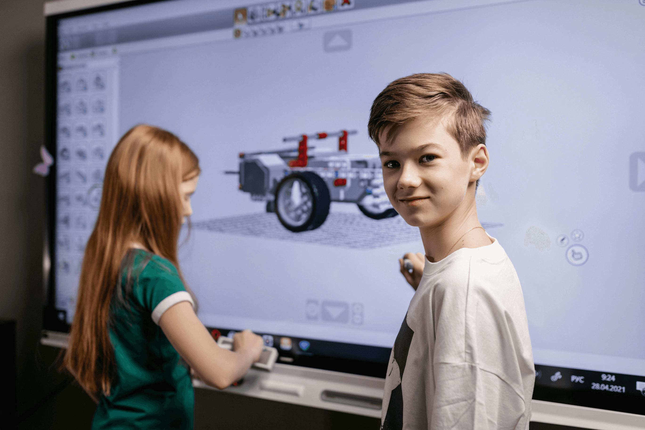 Two children are interacting with a large touchscreen displaying a 3D model of a vehicle, seemingly engaged in a STEM or educational design activity in a classroom setting.
