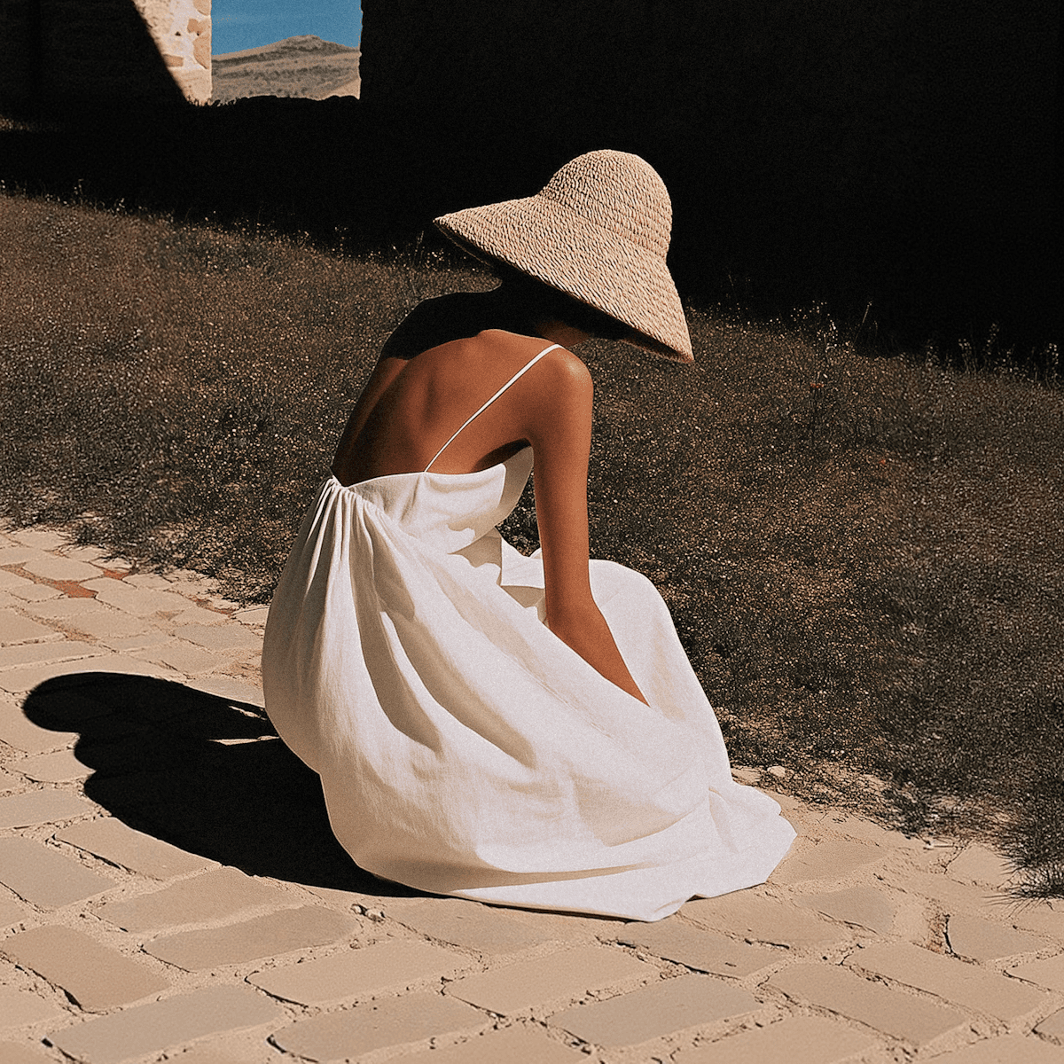 Woman in a white dress and wide straw hat sitting on a sunlit stone path in a rustic landscape