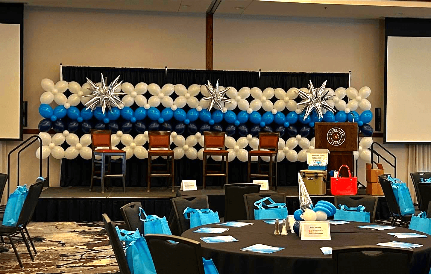 The image shows a conference room set up for an event, featuring a stage decorated with rows of blue, white, and silver balloons and flanked by large, metallic star-shaped balloons, surrounded by tables with blue gift bags.