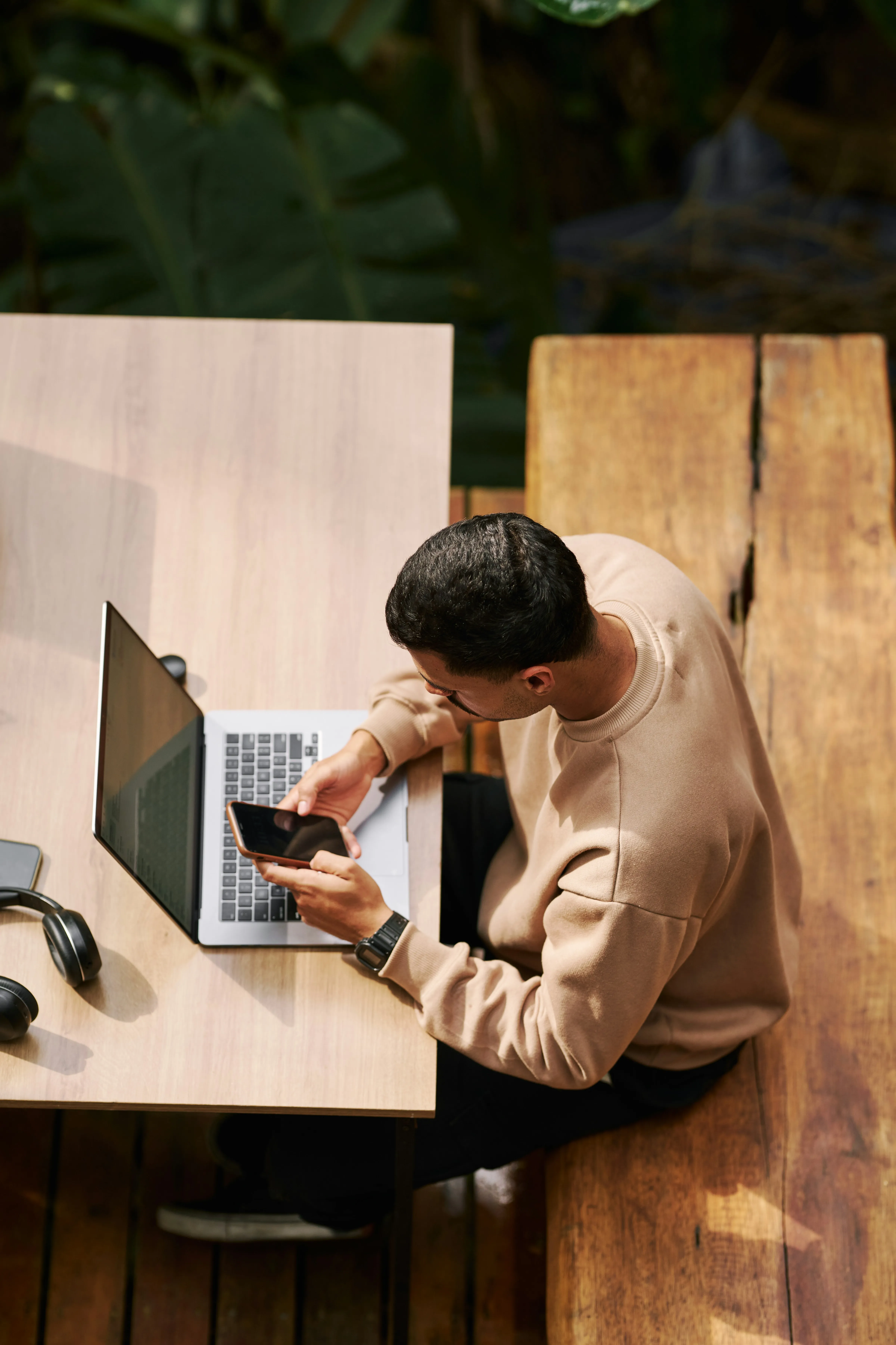 A person sits at a wooden table, working on a device with tools and a laptop nearby, outdoors among greenery.