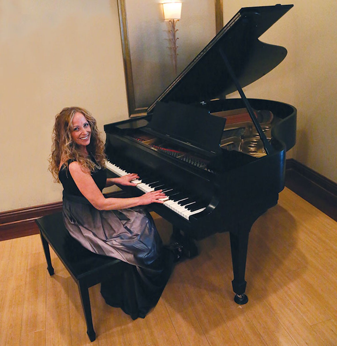 A smiling man with curly hair is sitting at a desk, looking at the camera, with a laptop and a lamp in the background.