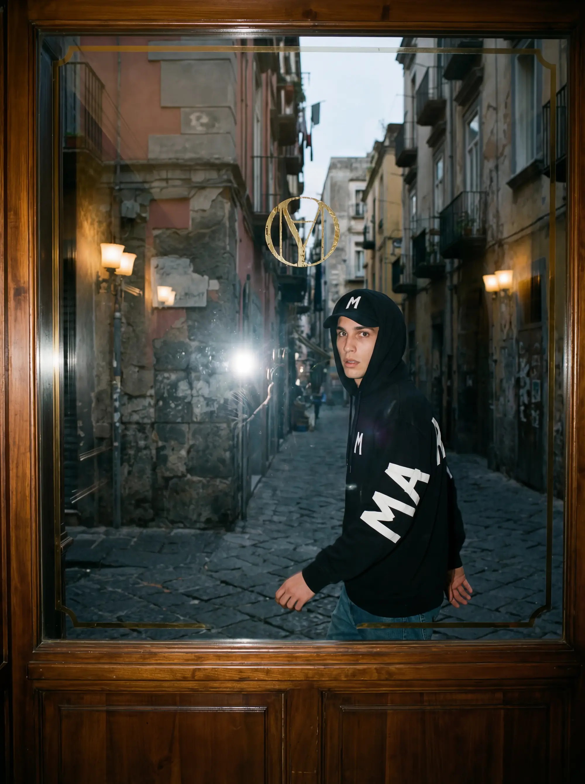 Male model wearing a black Maradona Official hoodie, reflected in an ornate glass door set in a narrow Napoli alleyway, with the brand monogram in gold above him