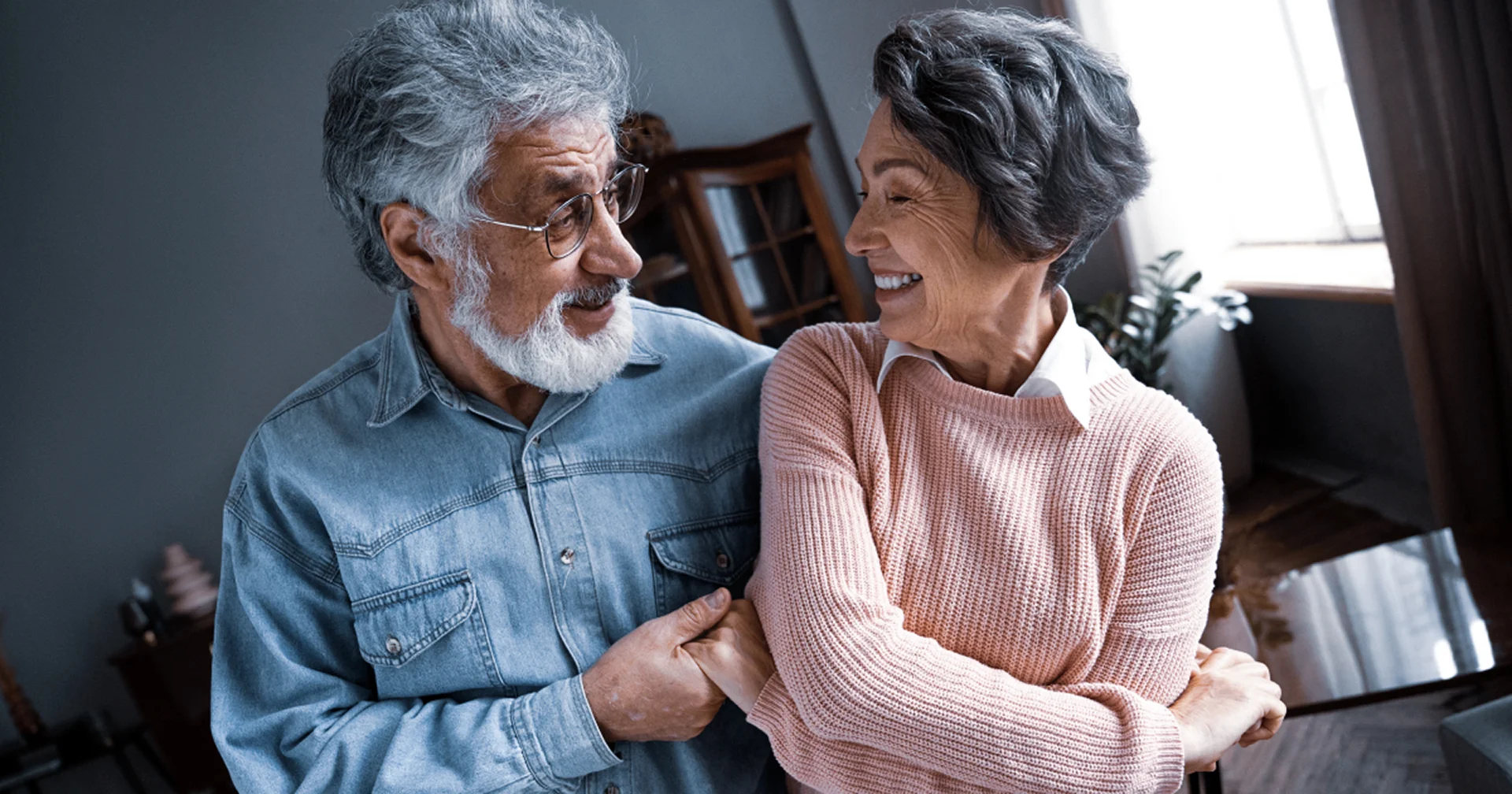 Casal de idosos sorridente e abraçado, com um homem de cabelos grisalhos e camisa jeans segurando o braço de uma mulher de suéter rosa claro enquanto se olham com carinho.