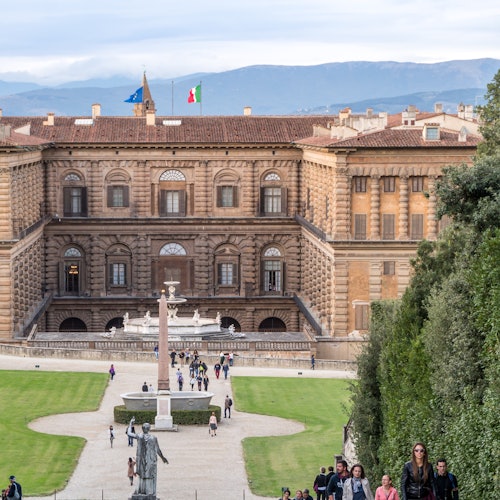 People walking in a garden leading to a large, historic building with two flags on top, with mountains in the background.