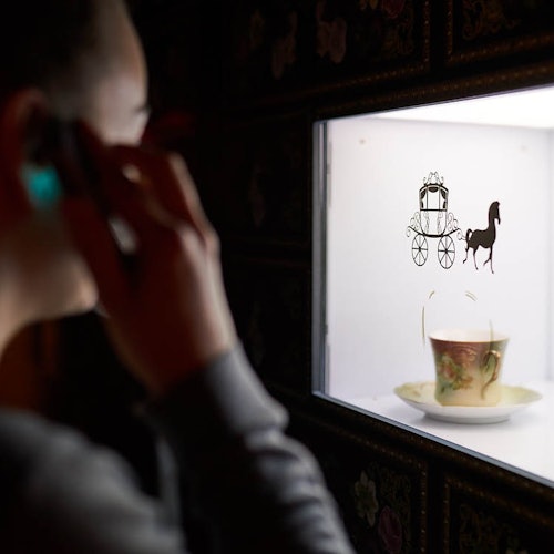 En person med en telefon tittar på en visningslåda med en tekopp och ett fat, samt en dekorativ silhuett av en häst och vagn på glaset.