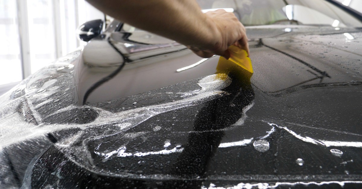 A hand is using a yellow squeegee to smooth clear protective film across the hood of a shiny black car.
