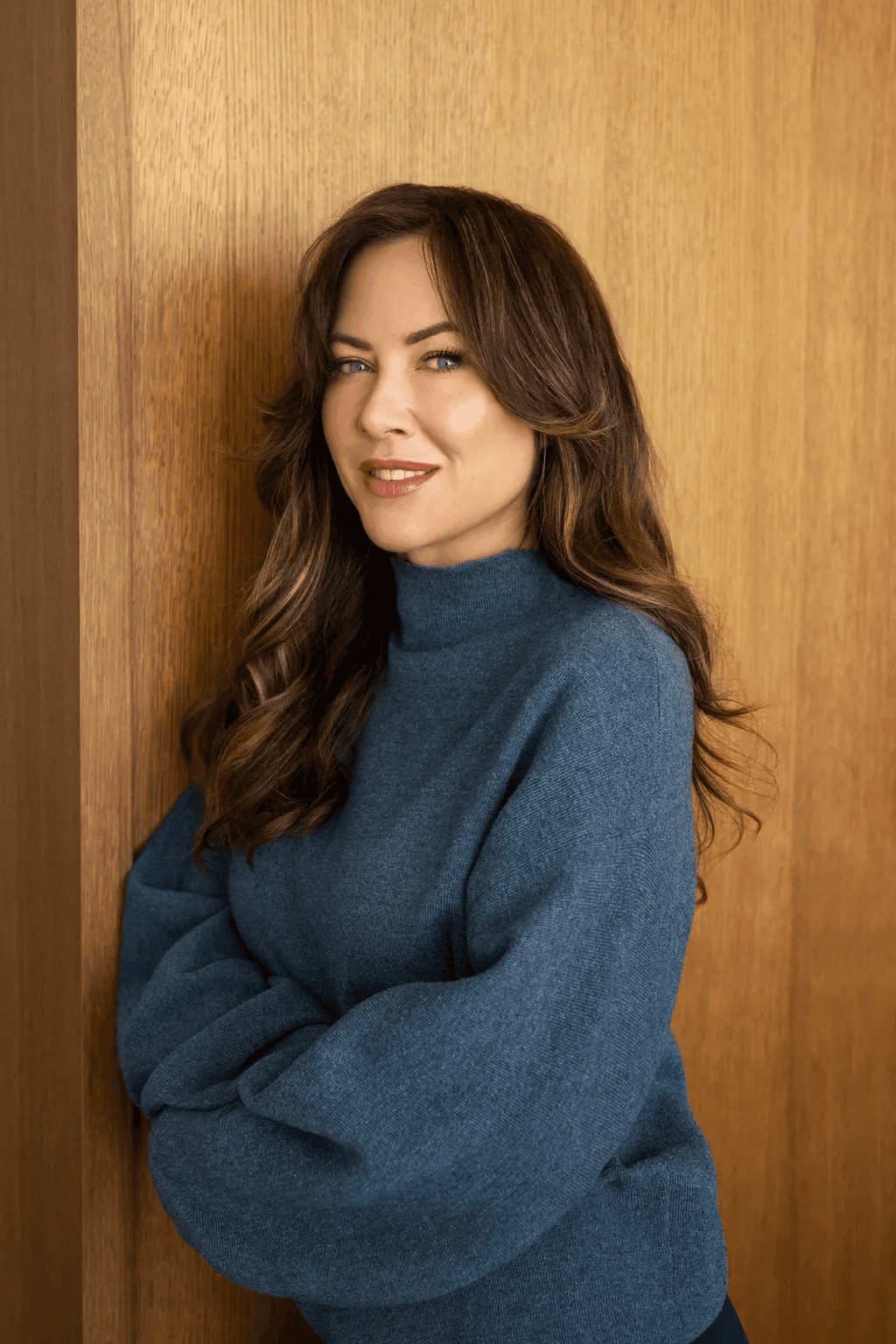 A person with long brown hair and a blue sweater leans casually against a wooden wall, smiling softly at the camera.