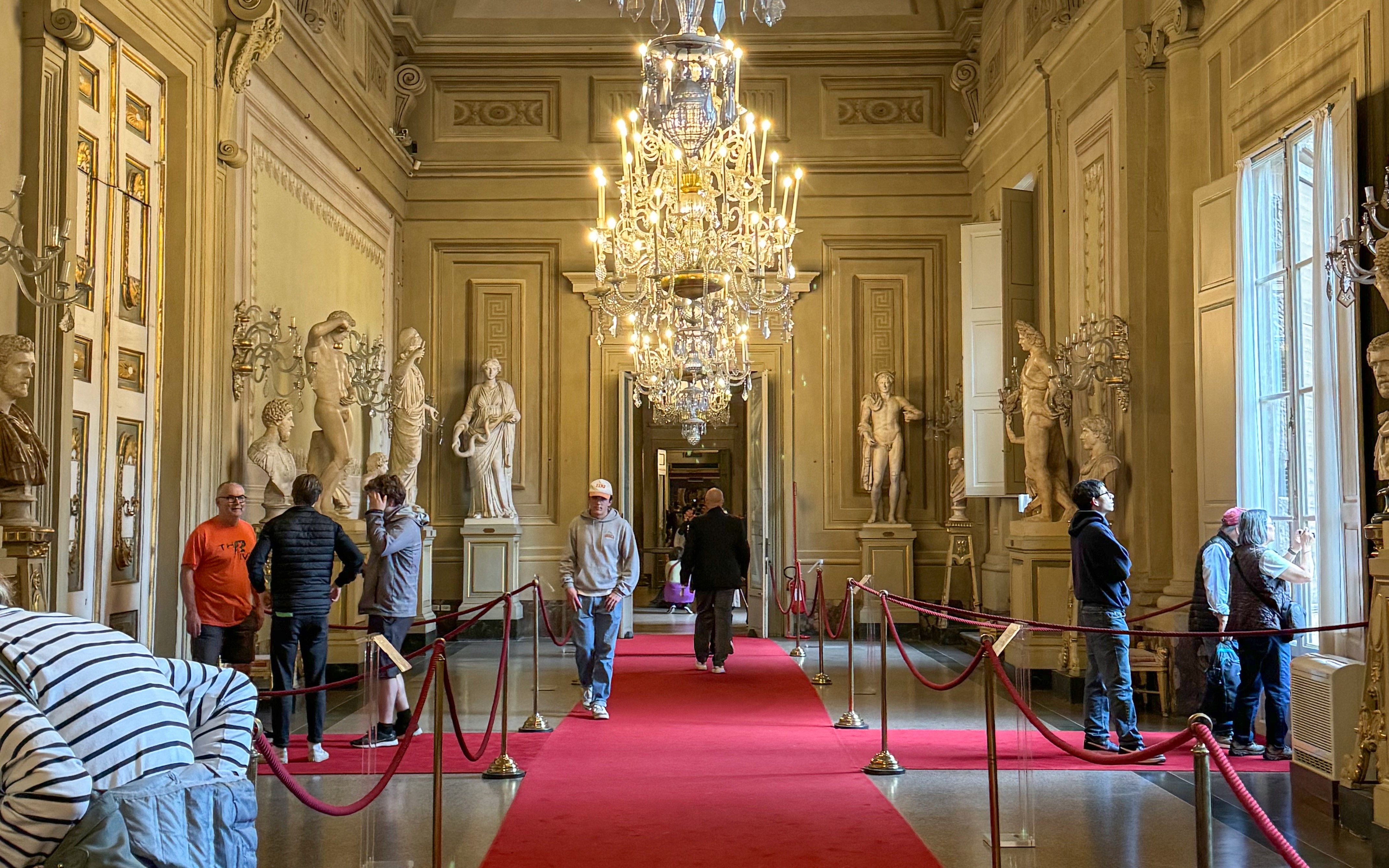 Visitors exploring statues and chandeliers in the Palatine Gallery, Pitti Palace, Florence.