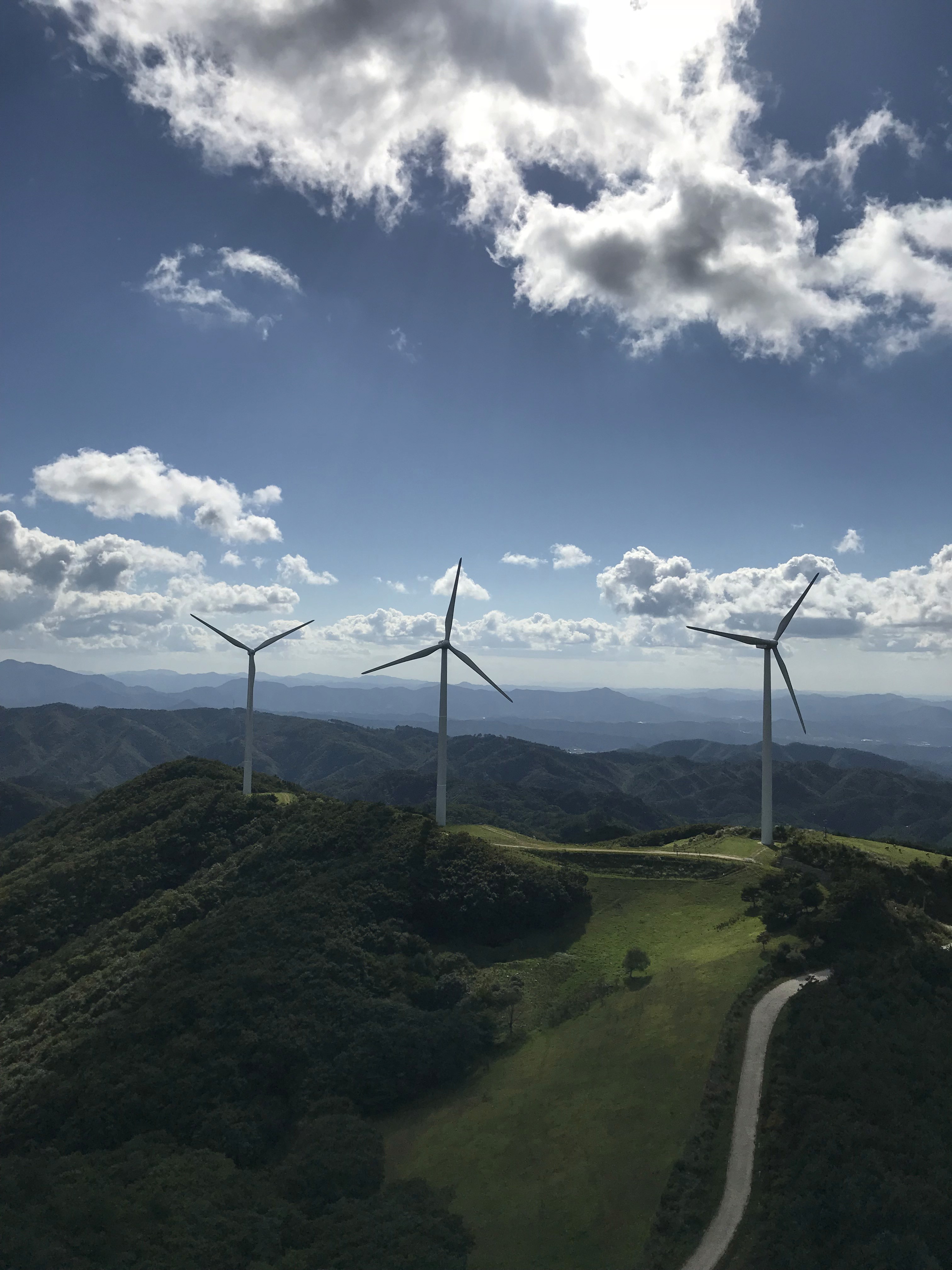 A scenic view of several wind turbines on a green hillside under a bright blue sky with scattered clouds.