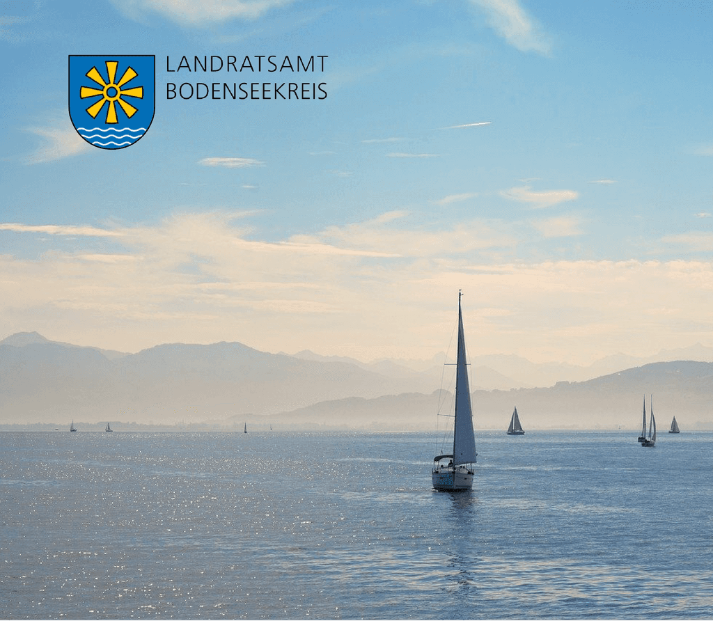 Mehrere Segelschiffe die sich auf dem Bodensee im Wasser befinden vor einem Bergpanorama an einem Sonnigen aber diesigen Tag, auf dem Bild befindet sich das Logo des Landratsamt Bodenseekreis