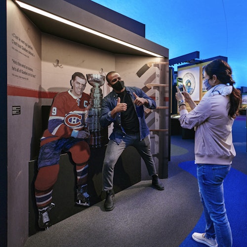 一个人站在冰球运动员的剪影和斯坦利杯复制品旁边，另一个人用智能手机拍照。